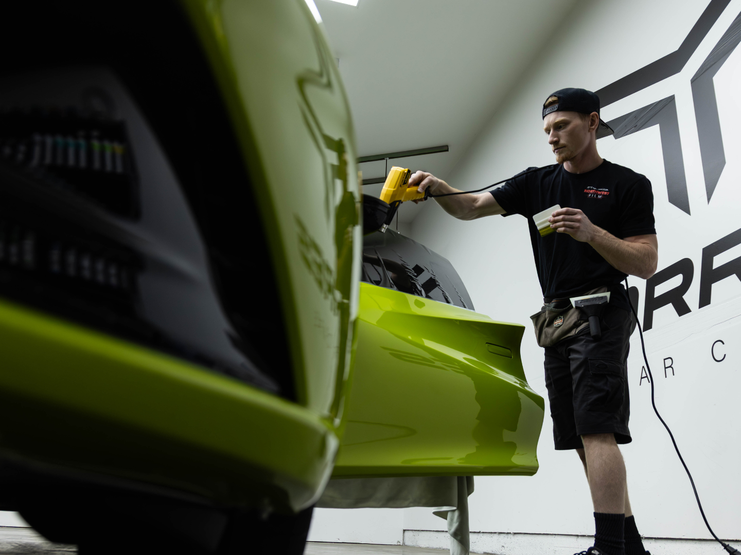 A man working on a green race car inside a garage or workshop, using a yellow tool, with a black and white logo on the wall behind him.