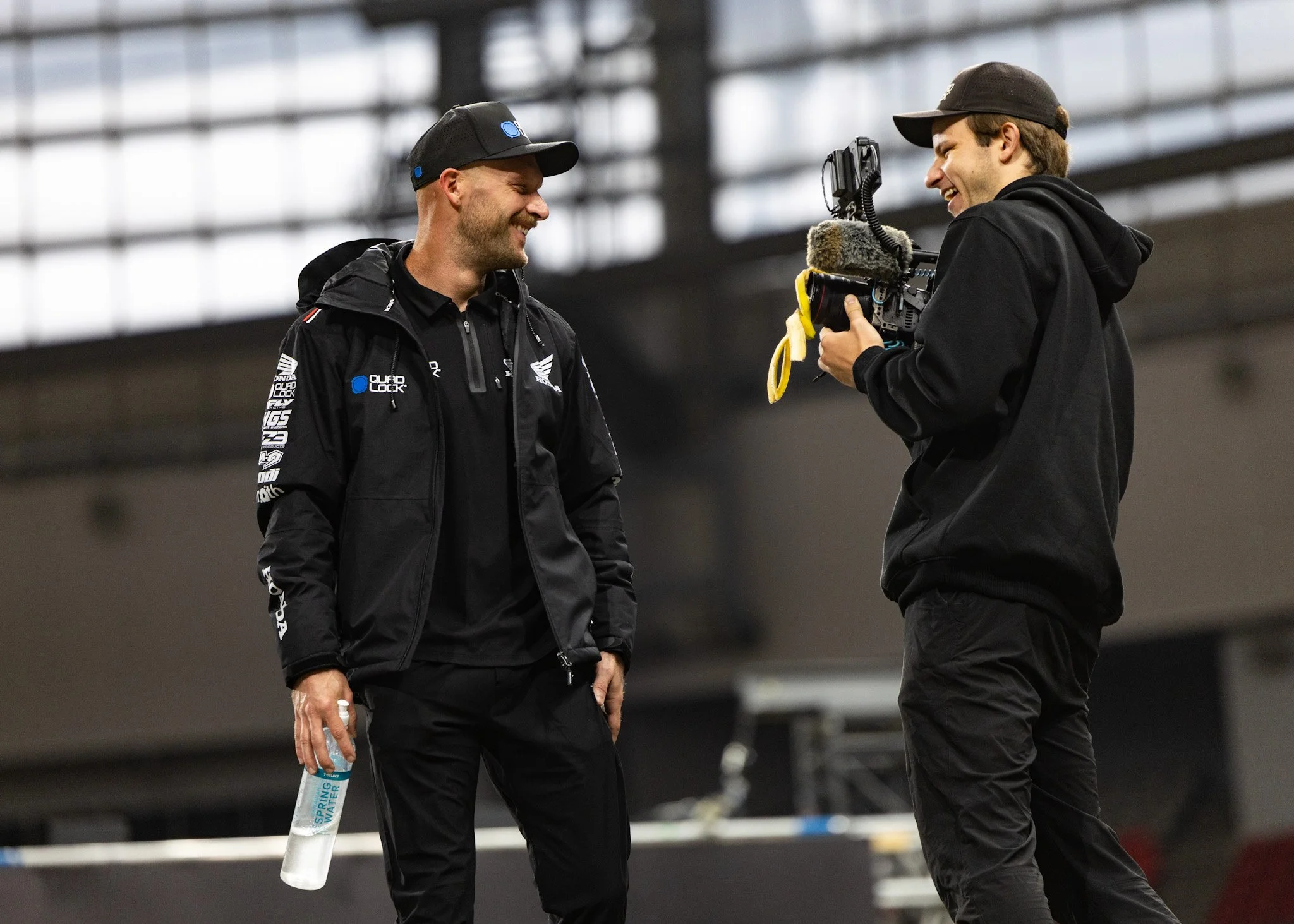 Two men standing and talking in an indoor sports facility or arena, both smiling and holding water bottles, one wearing a jacket with Honda and racing logos, the other in black attire, with a camera on a stabilizer or gimbal.
