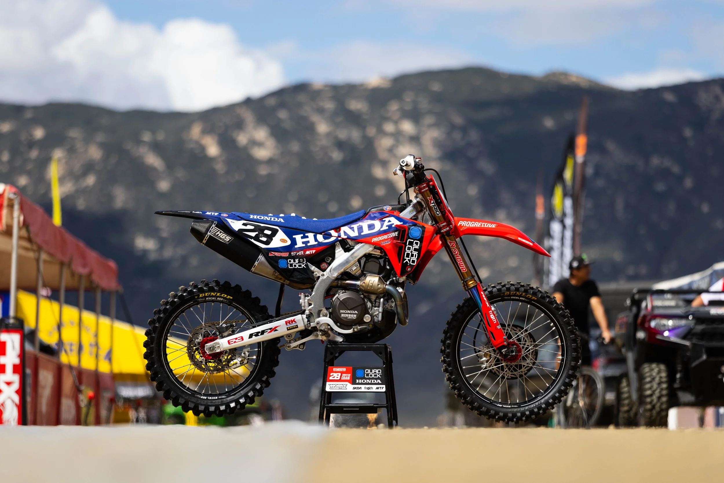 A motocross dirt bike on display at a racing event, with a mountain landscape and spectators in the background.