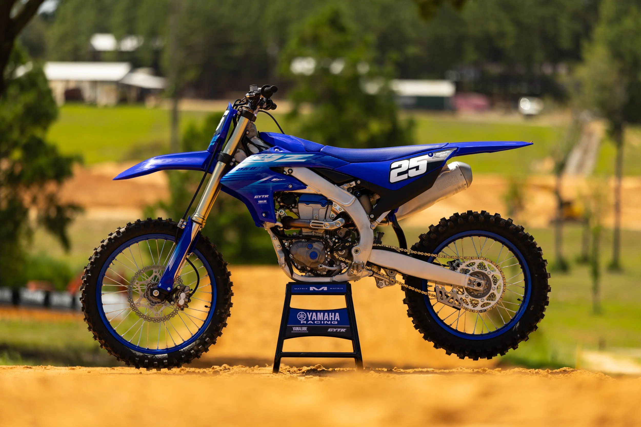 A blue Yamaha racing dirt bike displayed outdoors on a stand with a natural background of trees and grass.