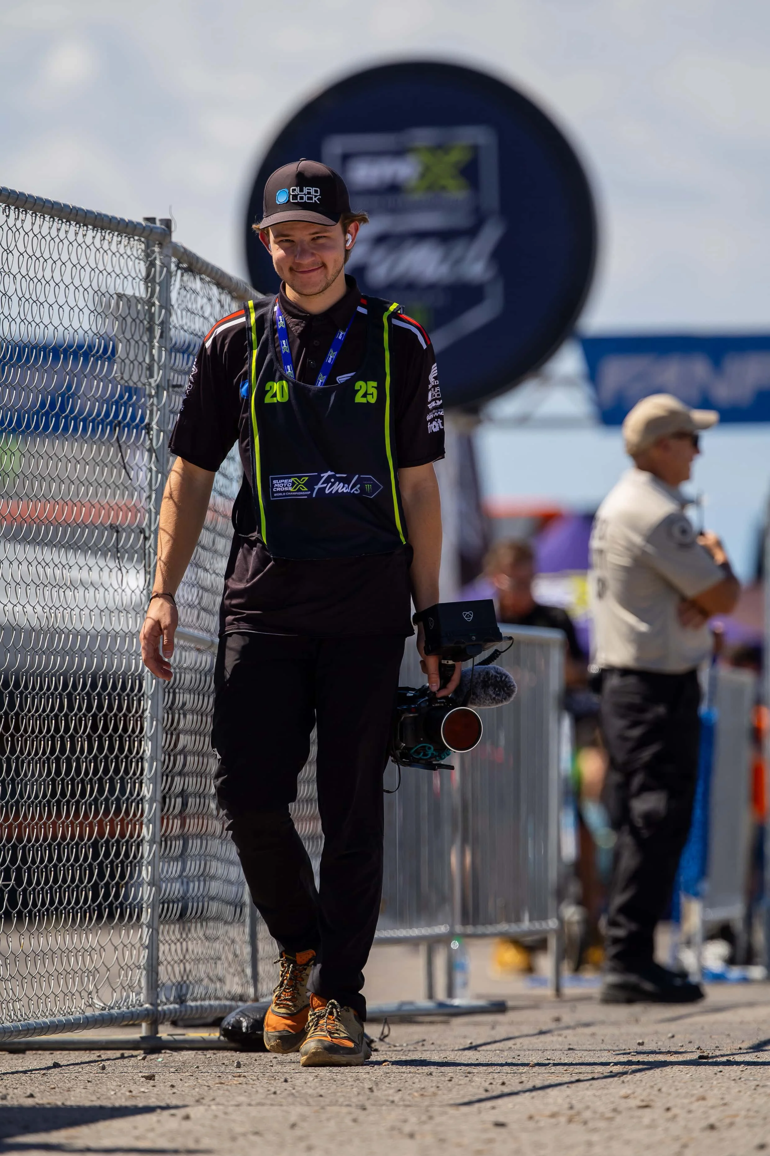 A young man walking along a chain-link fence with a camera in hand at a racing event. He is smiling, wearing a black cap, black racing uniform with yellow accents, and orange hiking shoes. In the background, there is a blurred person and a large sponsor banner.