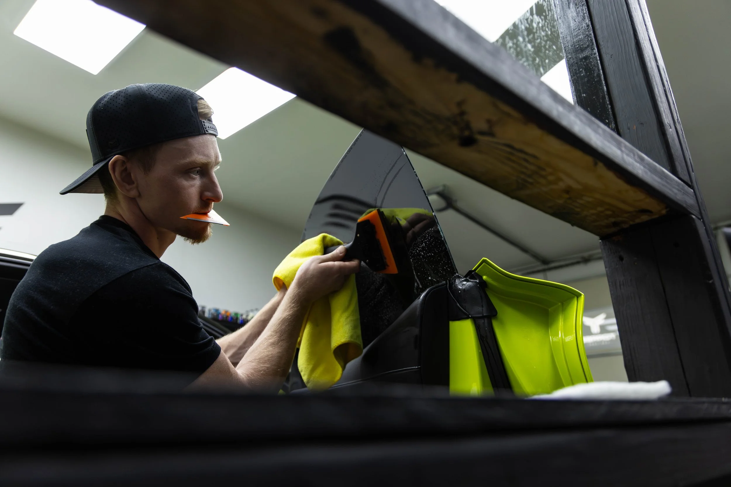 A man wearing a black cap and yellow gloves is working on a motorcycle inside a garage, with the focus on the motorcycle's windshield and the man's face.