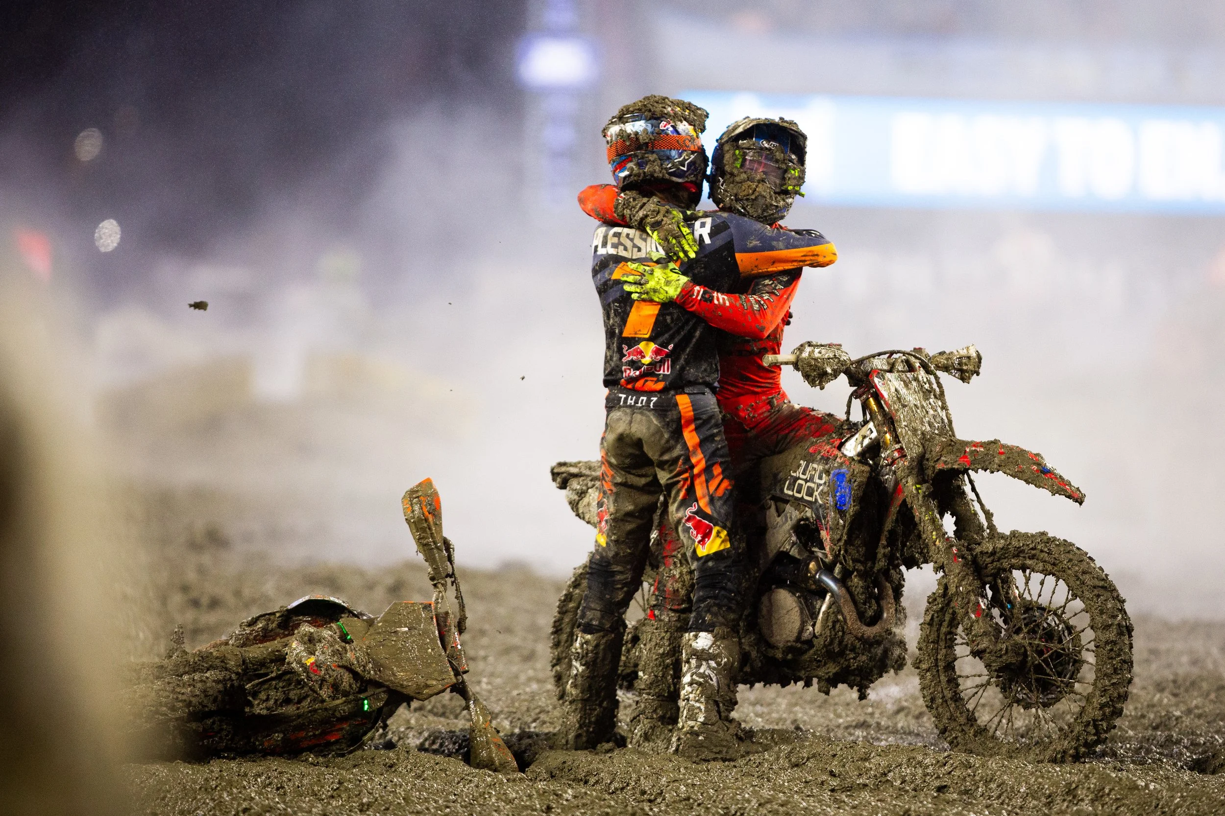Two dirt bike riders, covered in mud, embrace in a muddy environment during a race, with one rider resting on the other's shoulder while holding his bike.