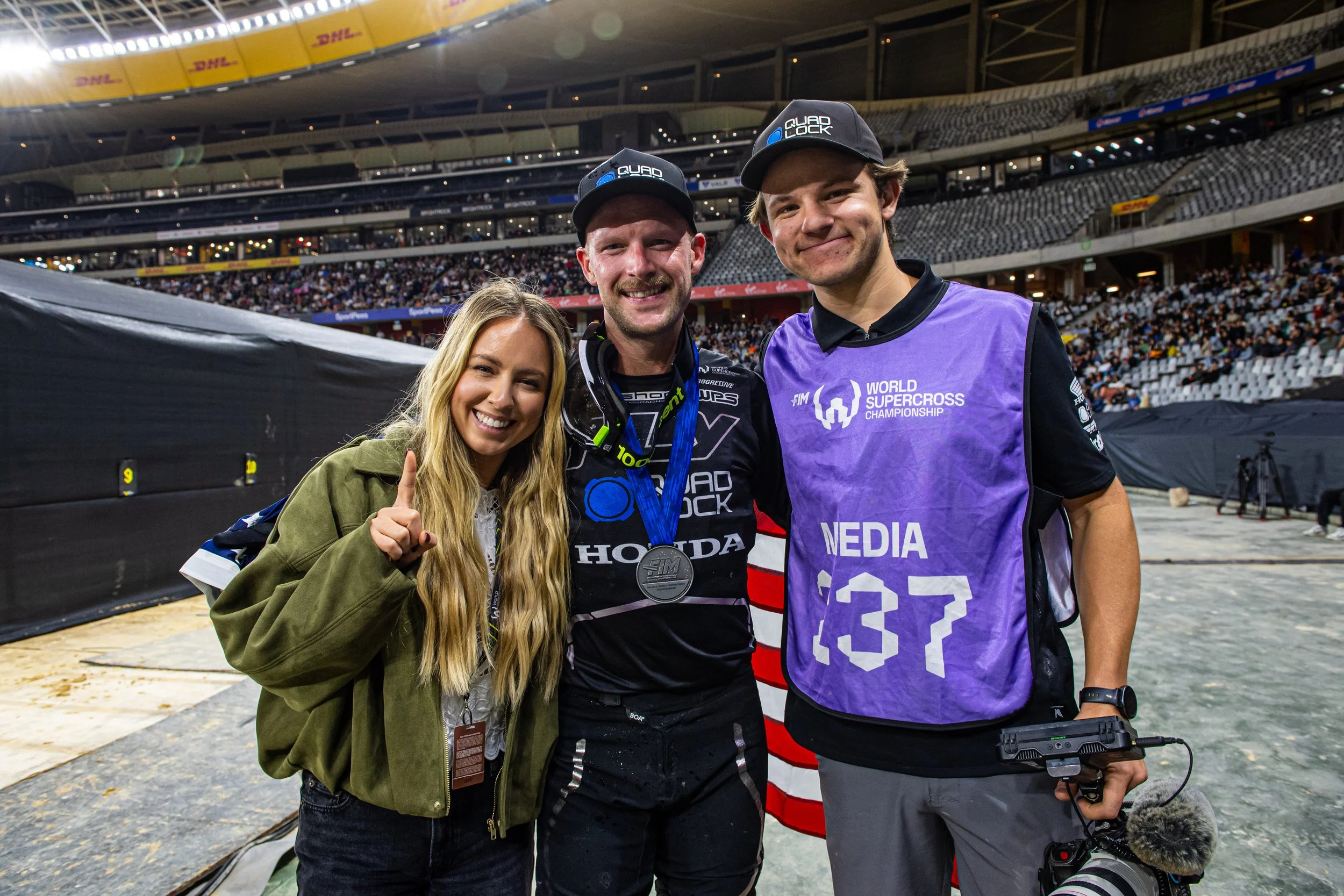 Three people smiling at a motocross event with a stadium in the background. The person in the middle wears a championship medal around their neck and a racing jersey, flanked by a woman and a man, both wearing caps and casual clothing.