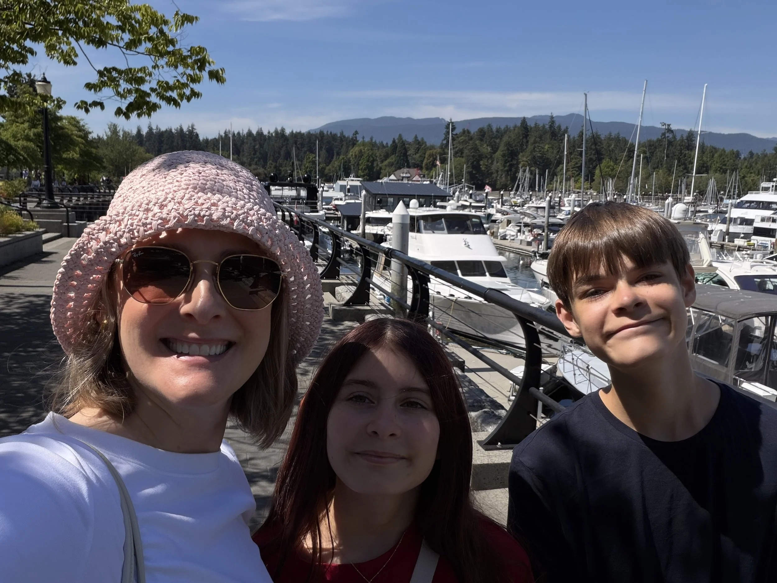 Courtney Charles and her children are taking a group selfie at a marina with boats and mountains in the background.
