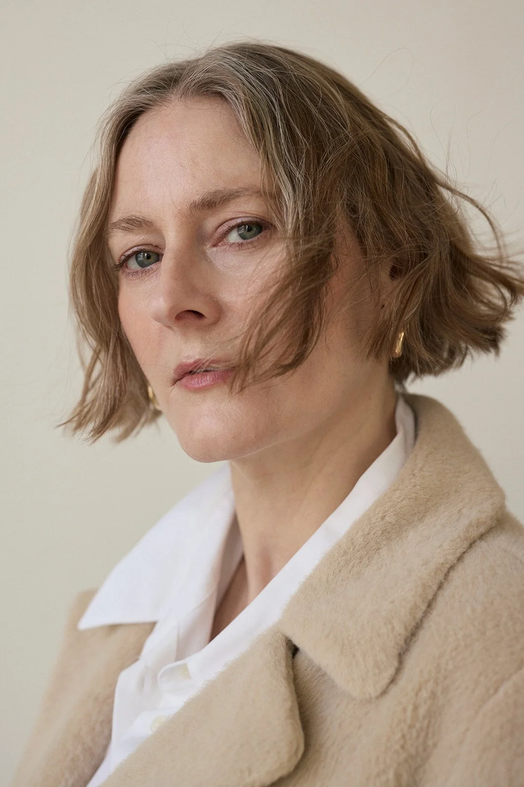 Close-up of Courtney Charles with wavy, shoulder-length hair, wearing a beige coat and white shirt, with minimal makeup.