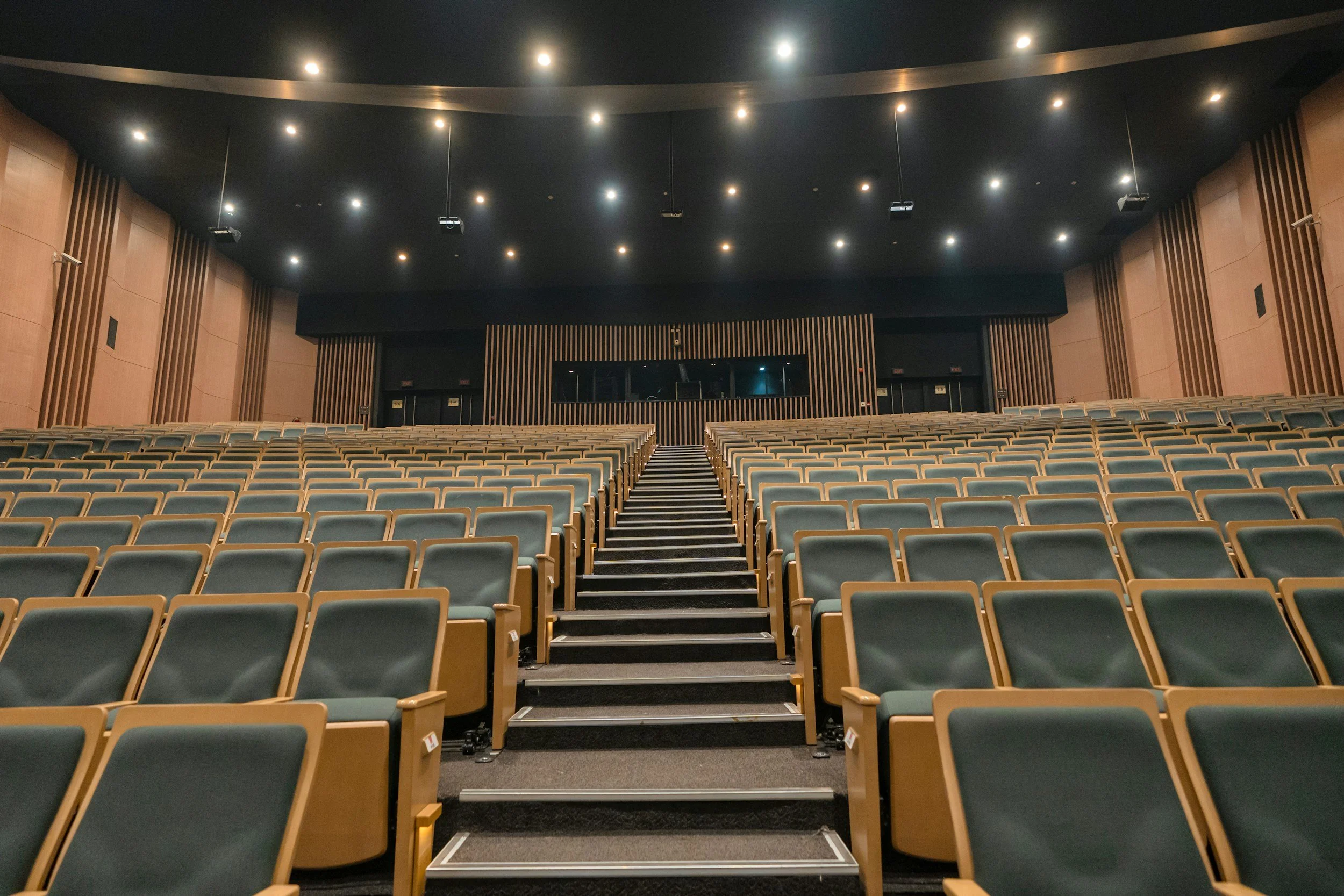 Empty auditorium with rows of green seats, a stage at the front, and overhead lights on a black ceiling.