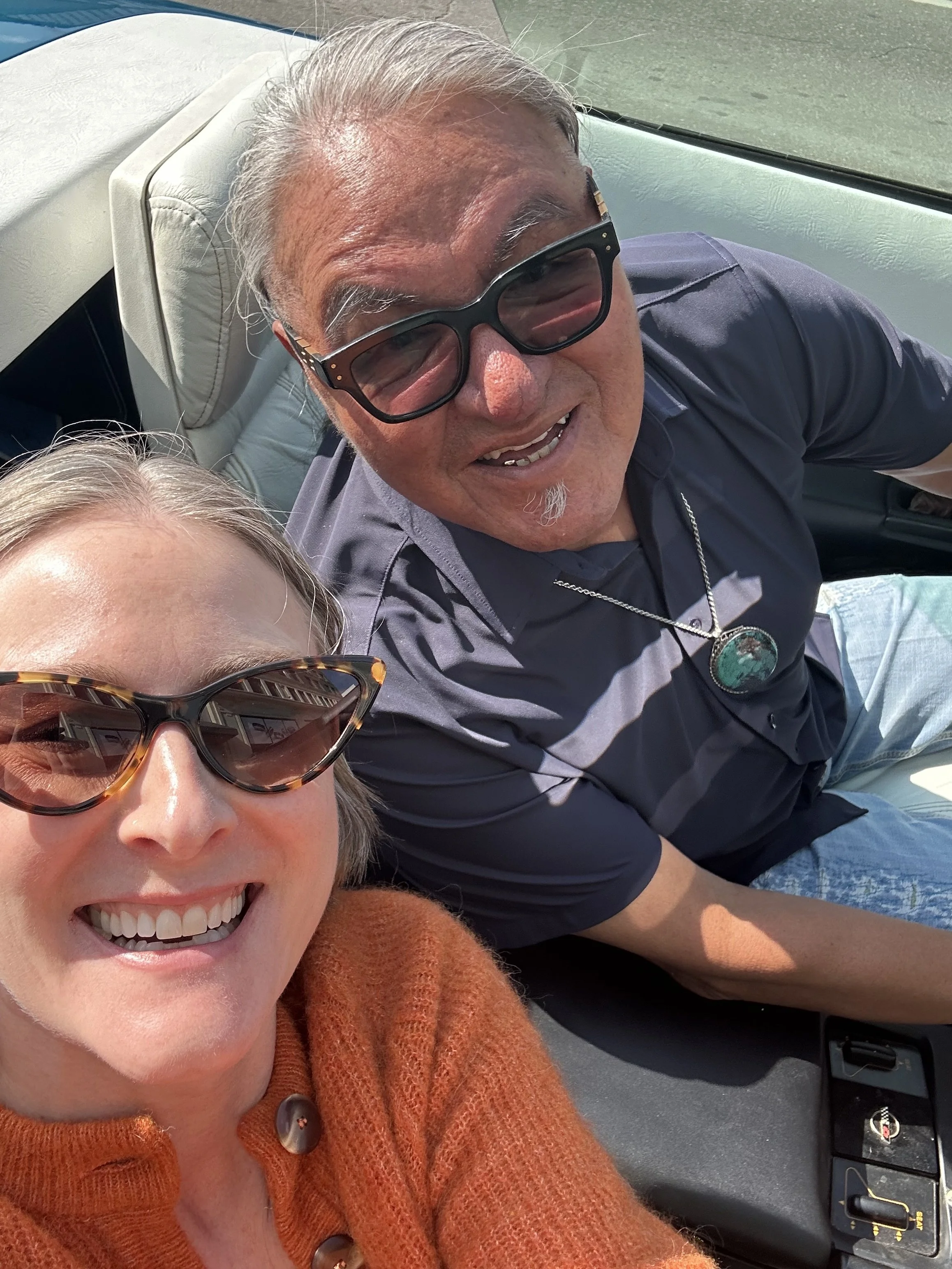 Close-up selfie of Courtney Charles with tortoise shell sunglasses, wearing an orange sweater, next to Harvey Thunderchild, wearing black sunglasses, sitting in a parked vehicle.