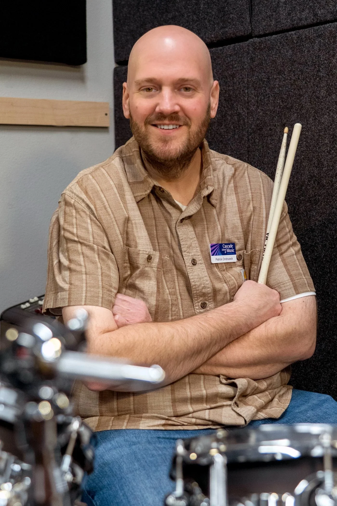 A man with a bald head, beard, and light skin sitting cross-armed at a table, smiling at the camera, in a recording studio or music room, holding drumsticks, wearing a plaid shirt and jeans.