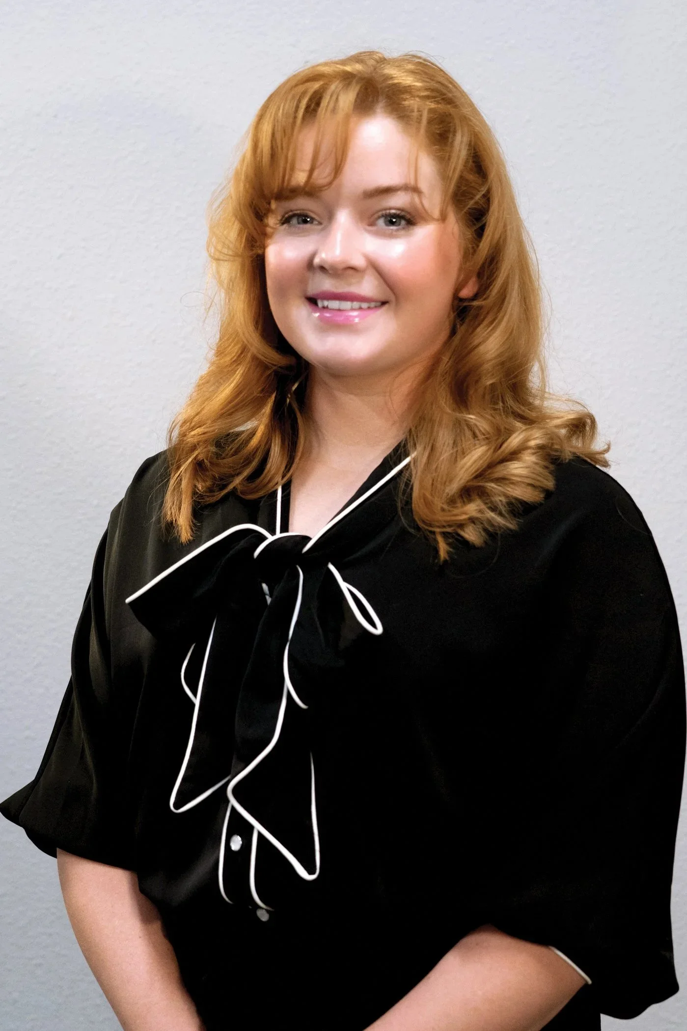 A woman with red hair smiling, wearing a black top with white piping and a bow detail.
