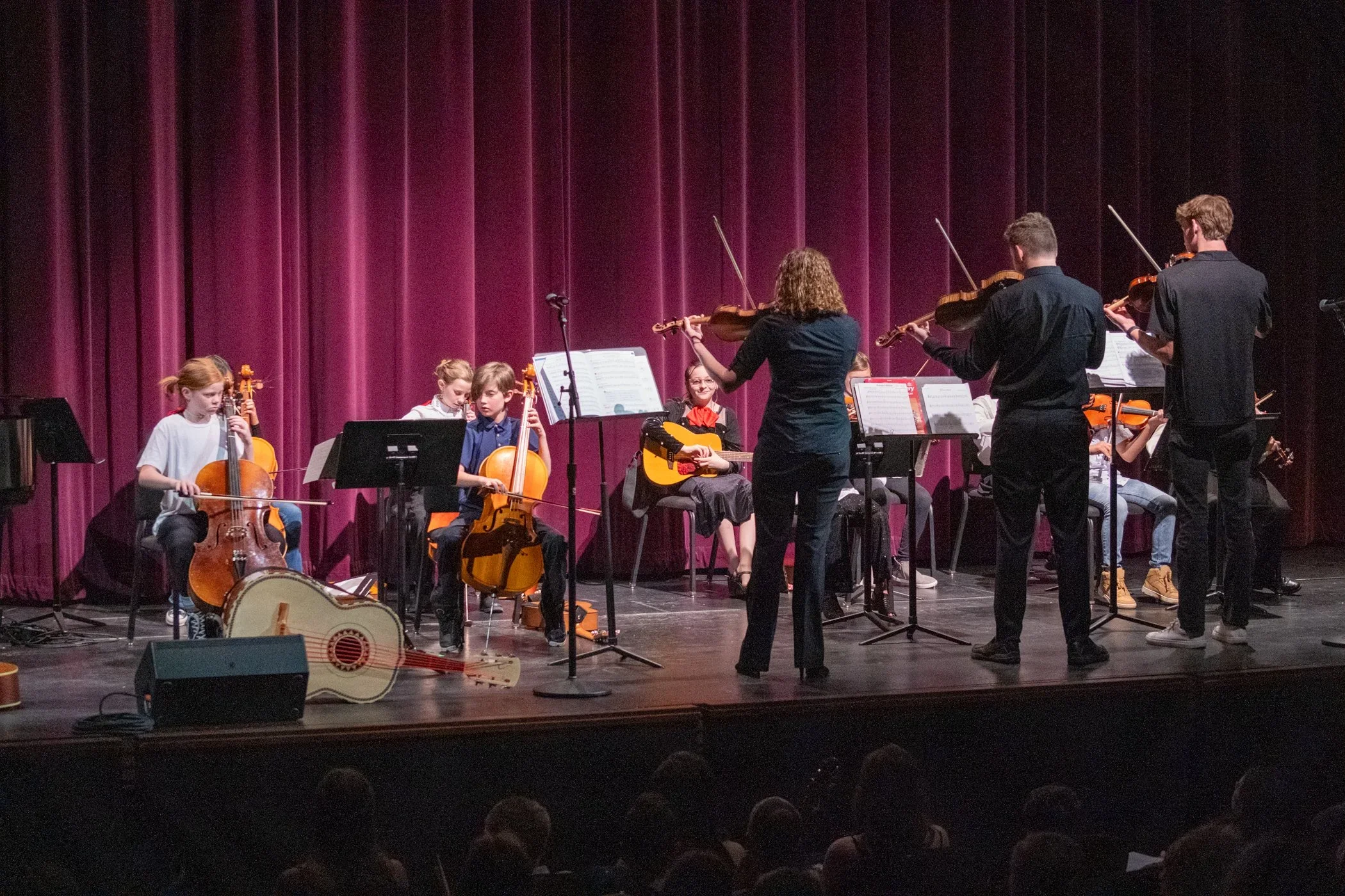 A group of young musicians performing on stage with instruments including violins, a guitar, and a cello, in front of a purple curtain, with an audience watching.