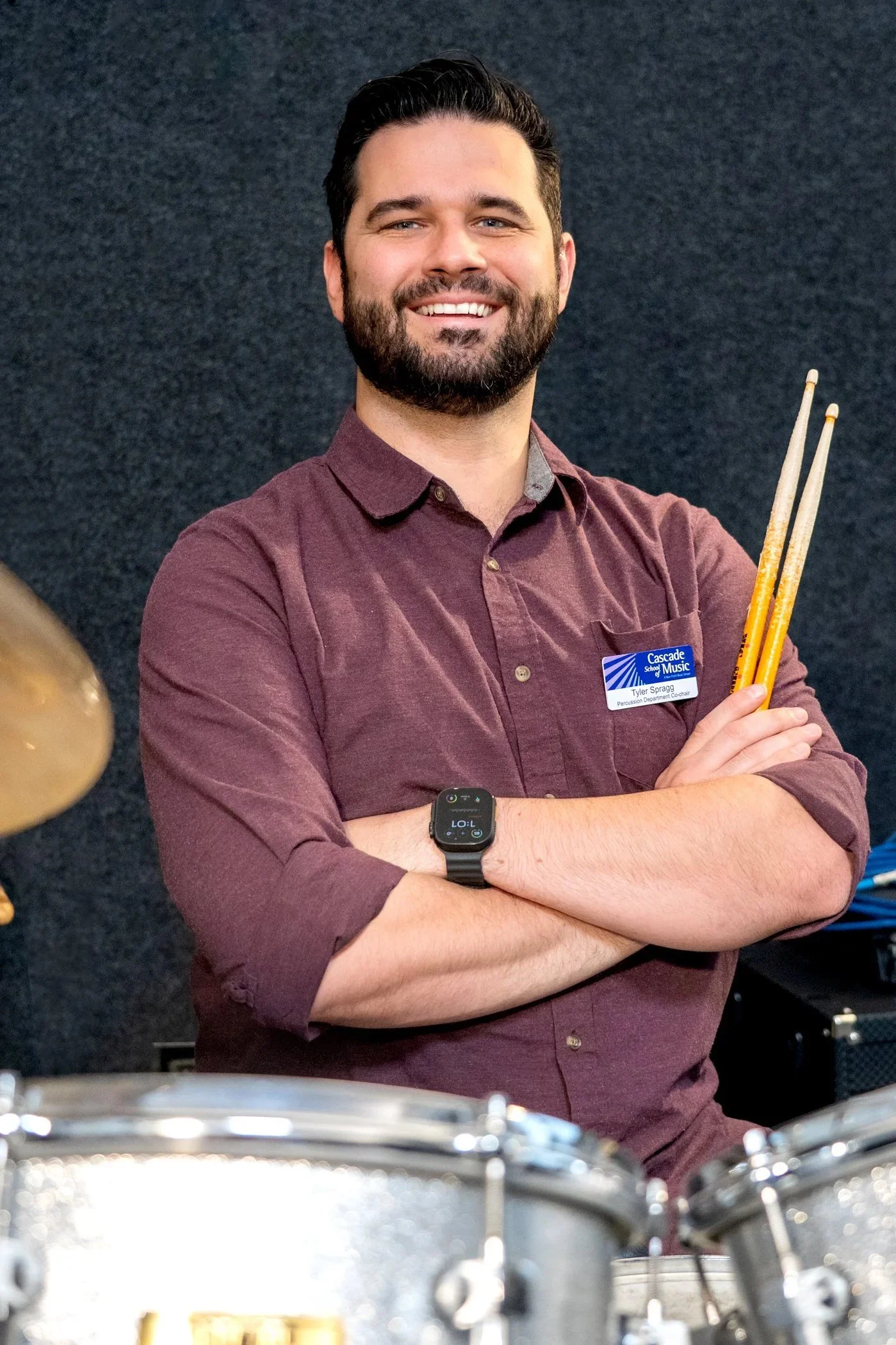 A smiling man with a beard wearing a maroon shirt and a name tag, holding drumsticks, standing in front of a dark background.