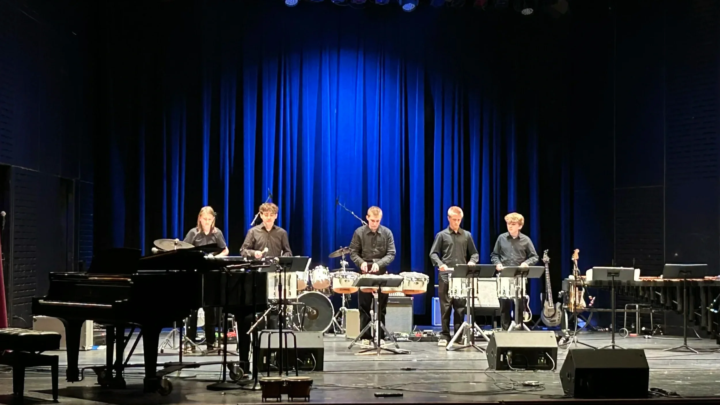 A group of musicians performing on stage with a blue curtain background, including a pianist, percussionists, and other instrumentalists.
