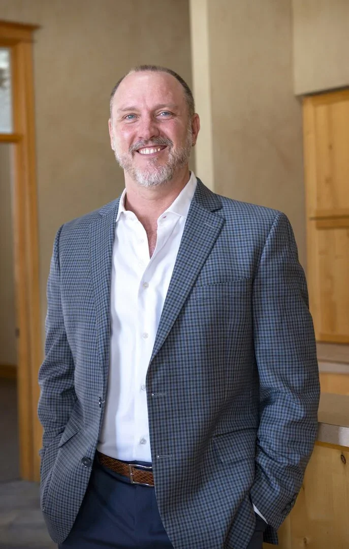 A smiling man with a beard and short hair wearing a plaid blazer and white shirt, standing indoors near wooden furniture.