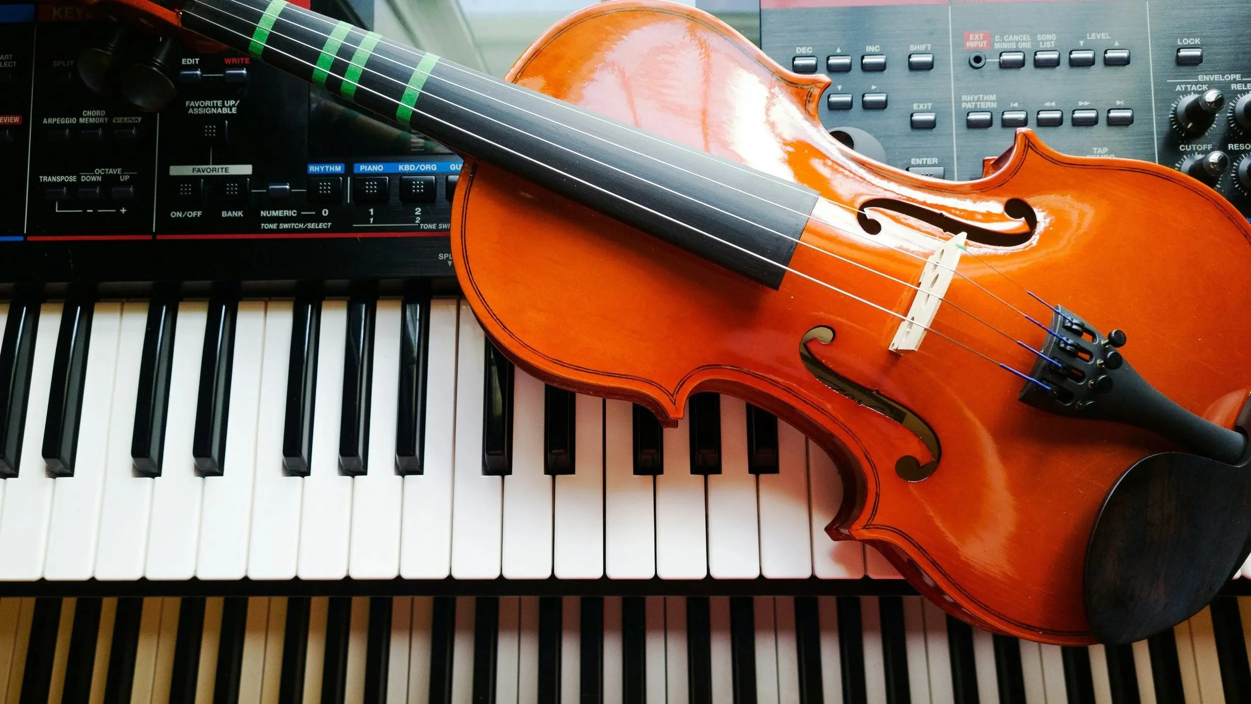 A musical setup featuring a keyboard, a violin resting on the keyboard, and a synthesizer in the background.