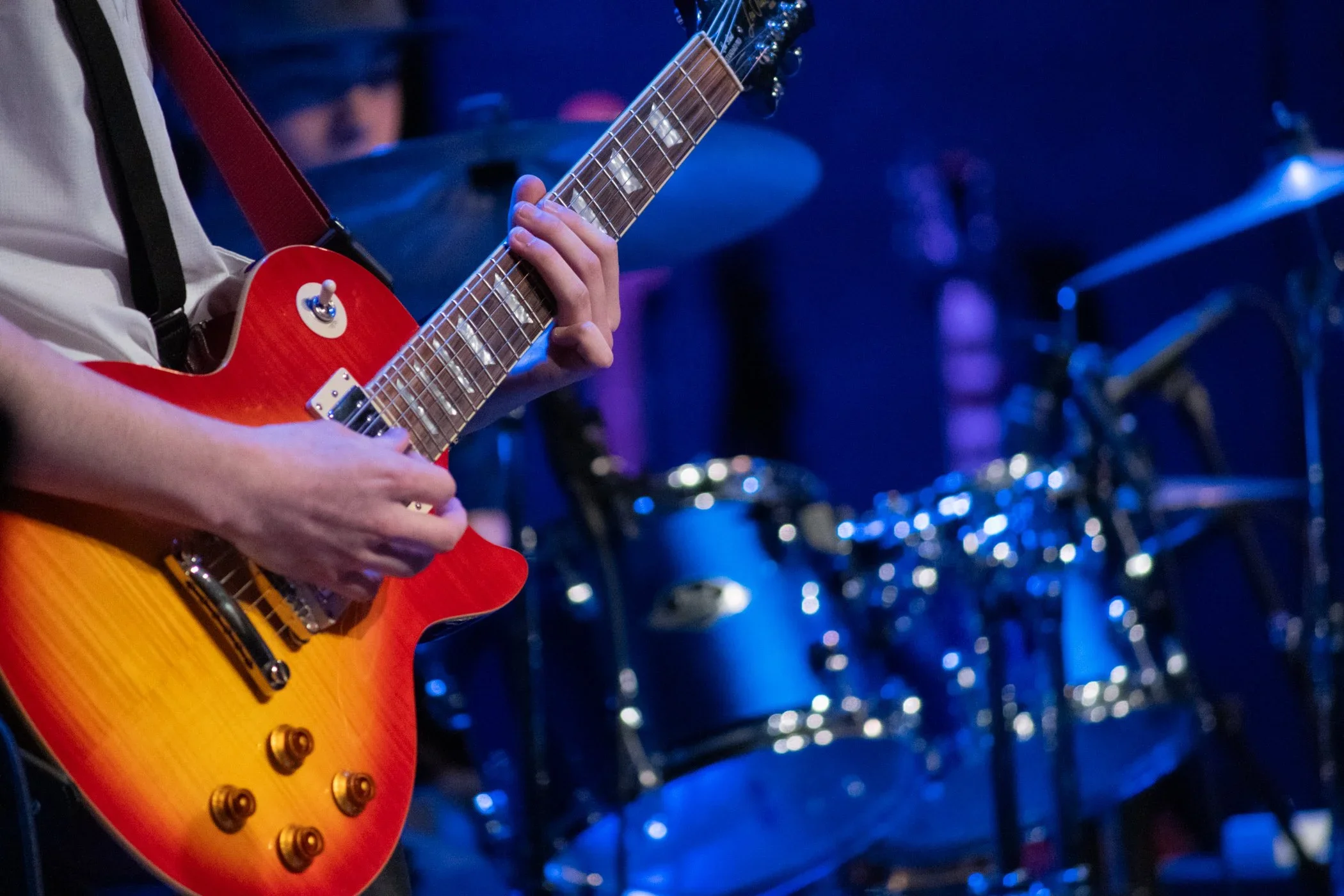 Person playing an electric guitar with a red and yellow sunburst finish, wearing a white shirt, against a dark and blurry background with blue lighting.