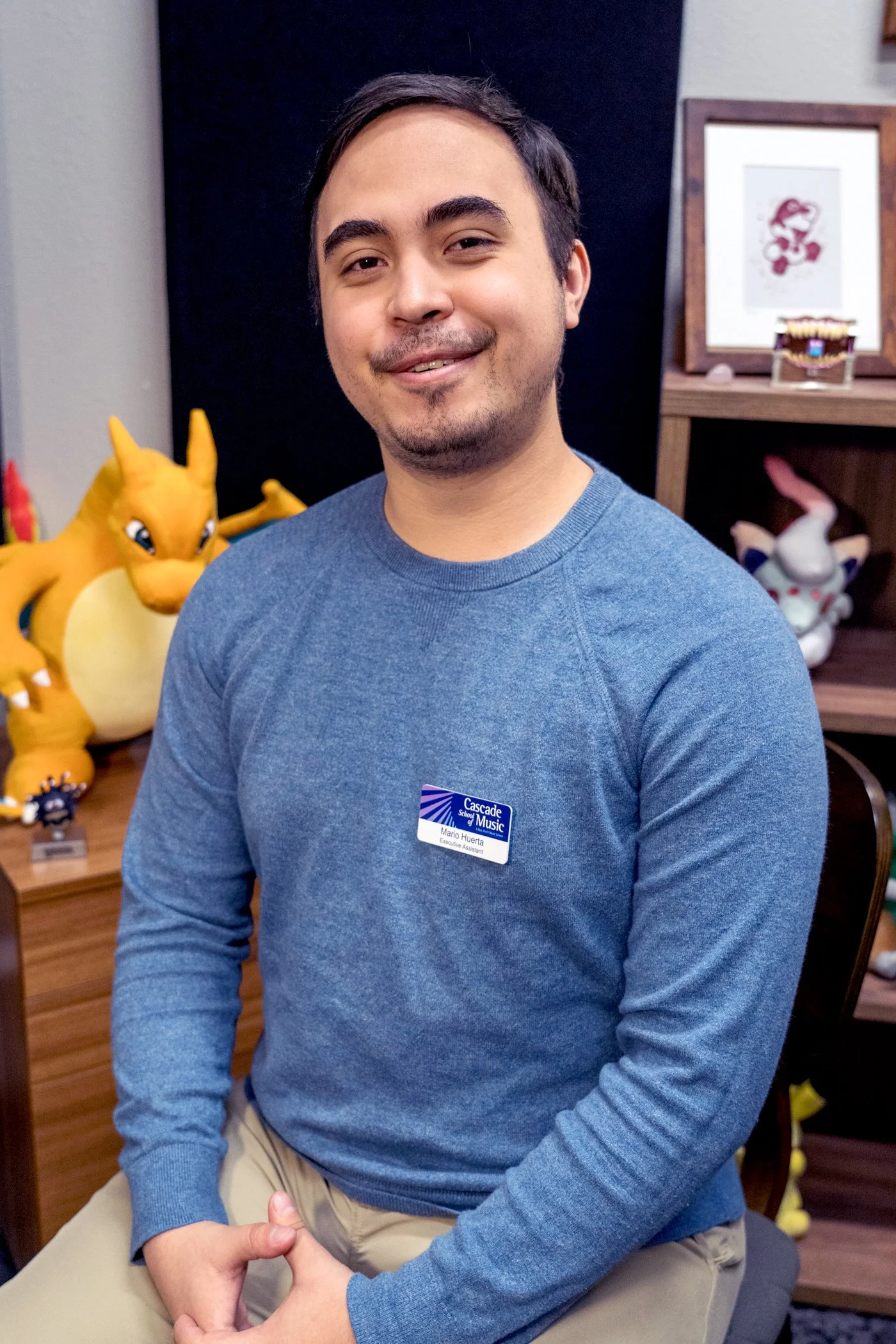 A young man with short dark hair, a light beard, and a friendly smile, sitting indoors with a group of plush toys and framed artwork in the background.
