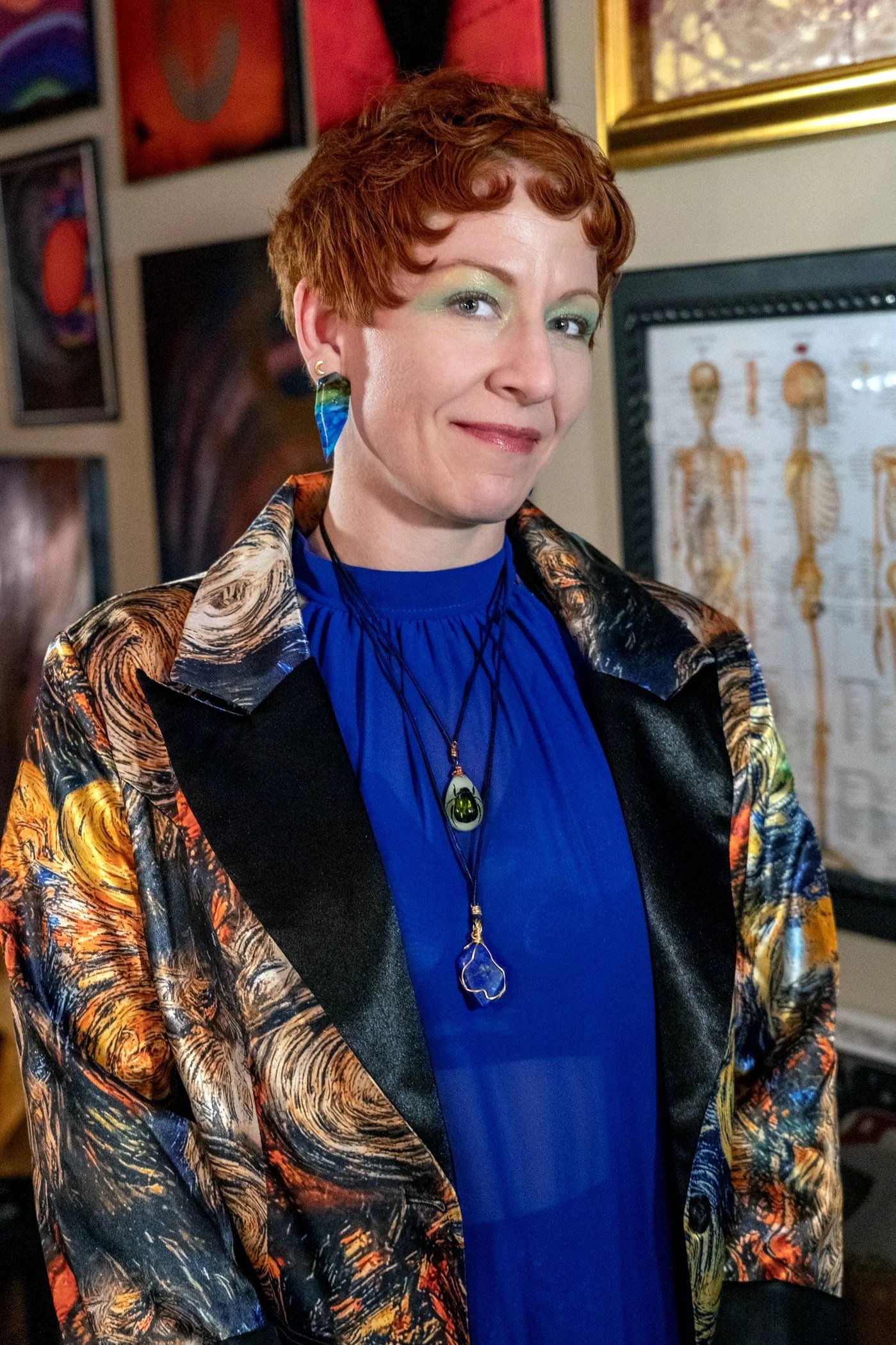 A woman with short red hair and light makeup wearing a vibrant multicolored jacket, a blue top, and a necklace with a large green pendant, standing indoors with framed artwork and a chart of human skeletons on the wall behind her.