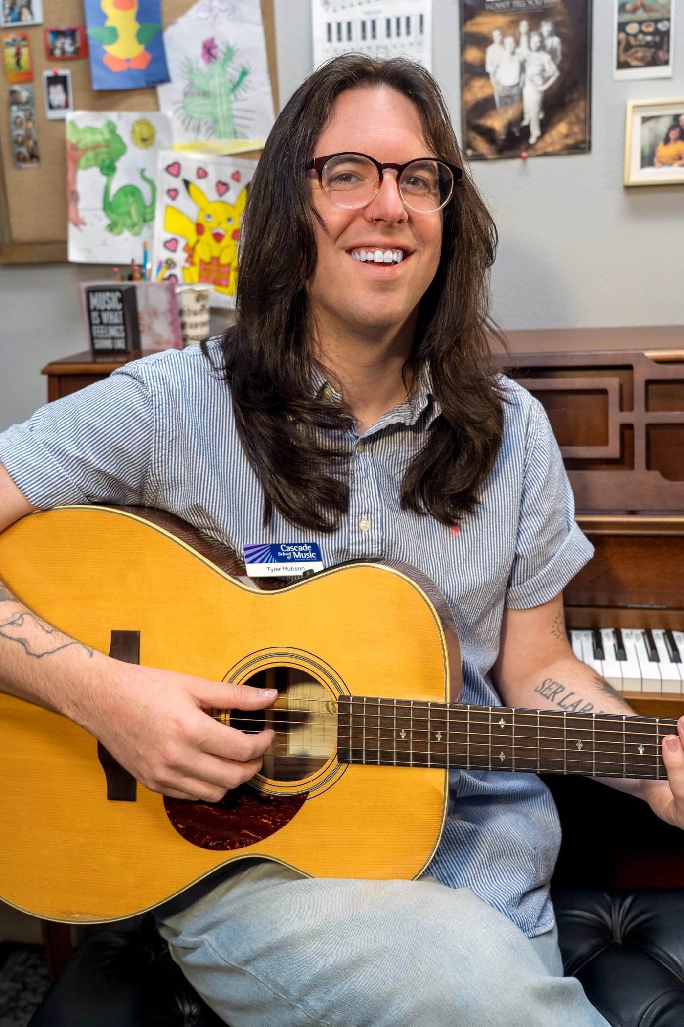 A person with long dark hair, glasses, and a light blue striped shirt playing an acoustic guitar. The background features colorful children's artwork, photographs, and posters on a wall, and a piano keyboard is partially visible.