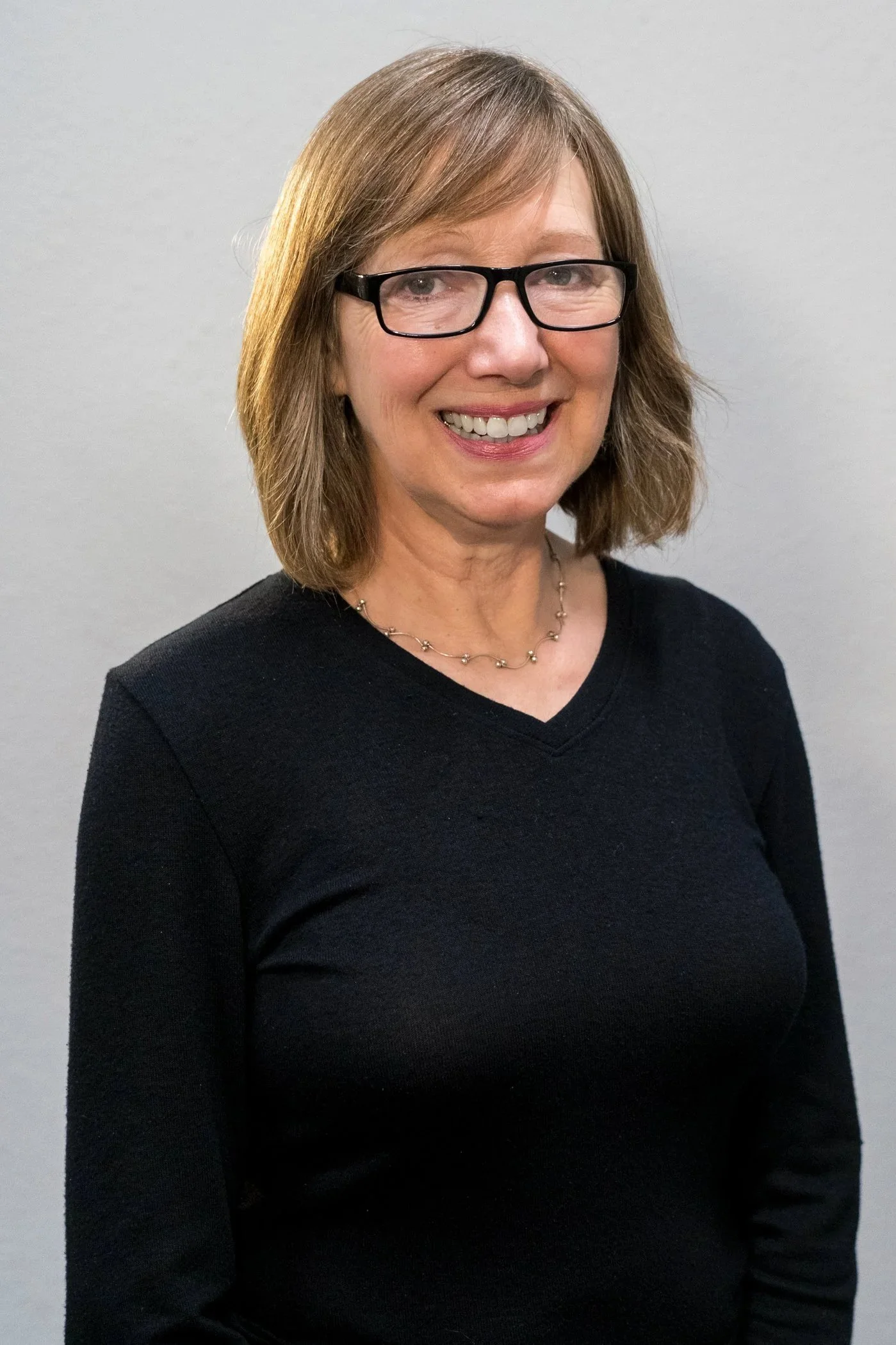 A woman with shoulder-length brown hair, wearing black glasses, a black top, and a gold necklace, smiling against a plain gray background.