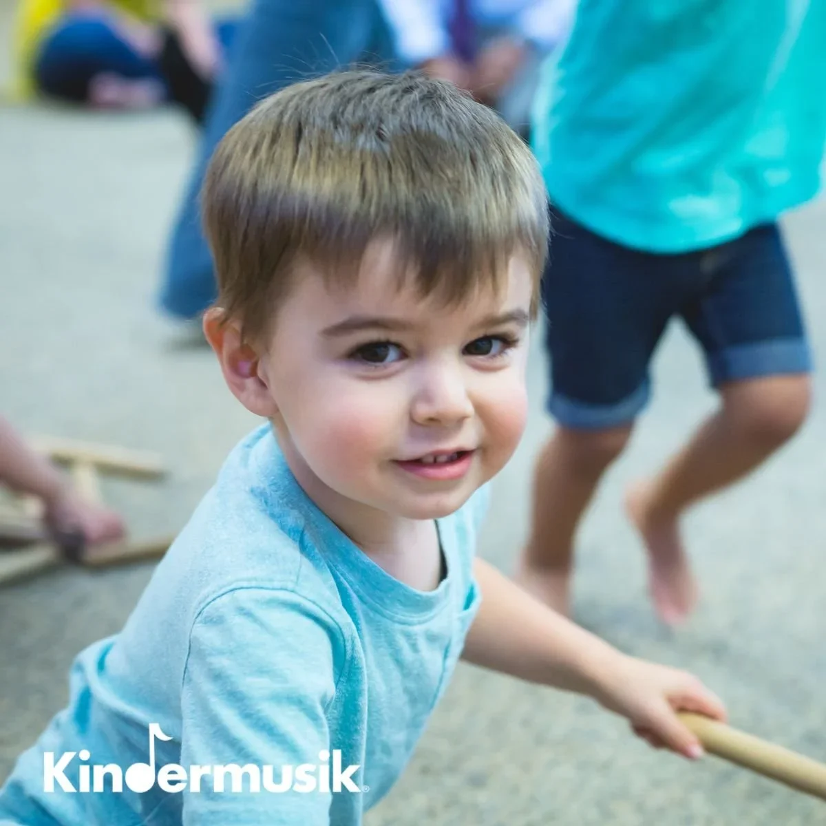 Young boy with brown hair and blue eyes looking at camera, holding a wooden stick, in a classroom or play area with other children in the background.