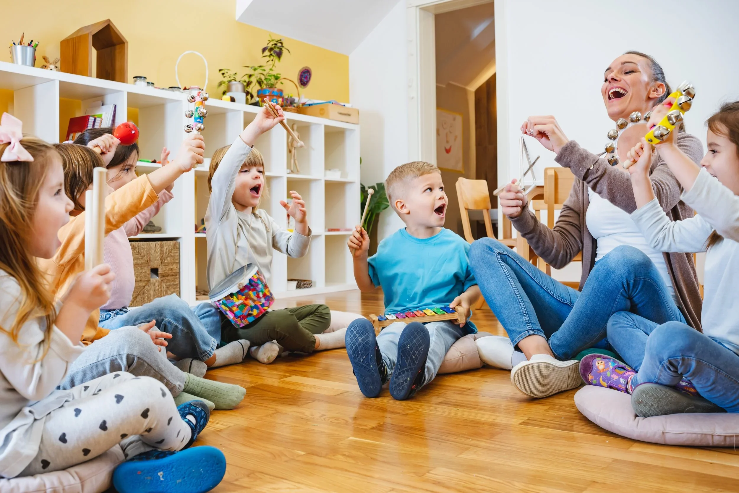 A group of children and a woman playing musical instruments and singing together in a cozy room with wooden flooring and white shelves, filled with books and plants.