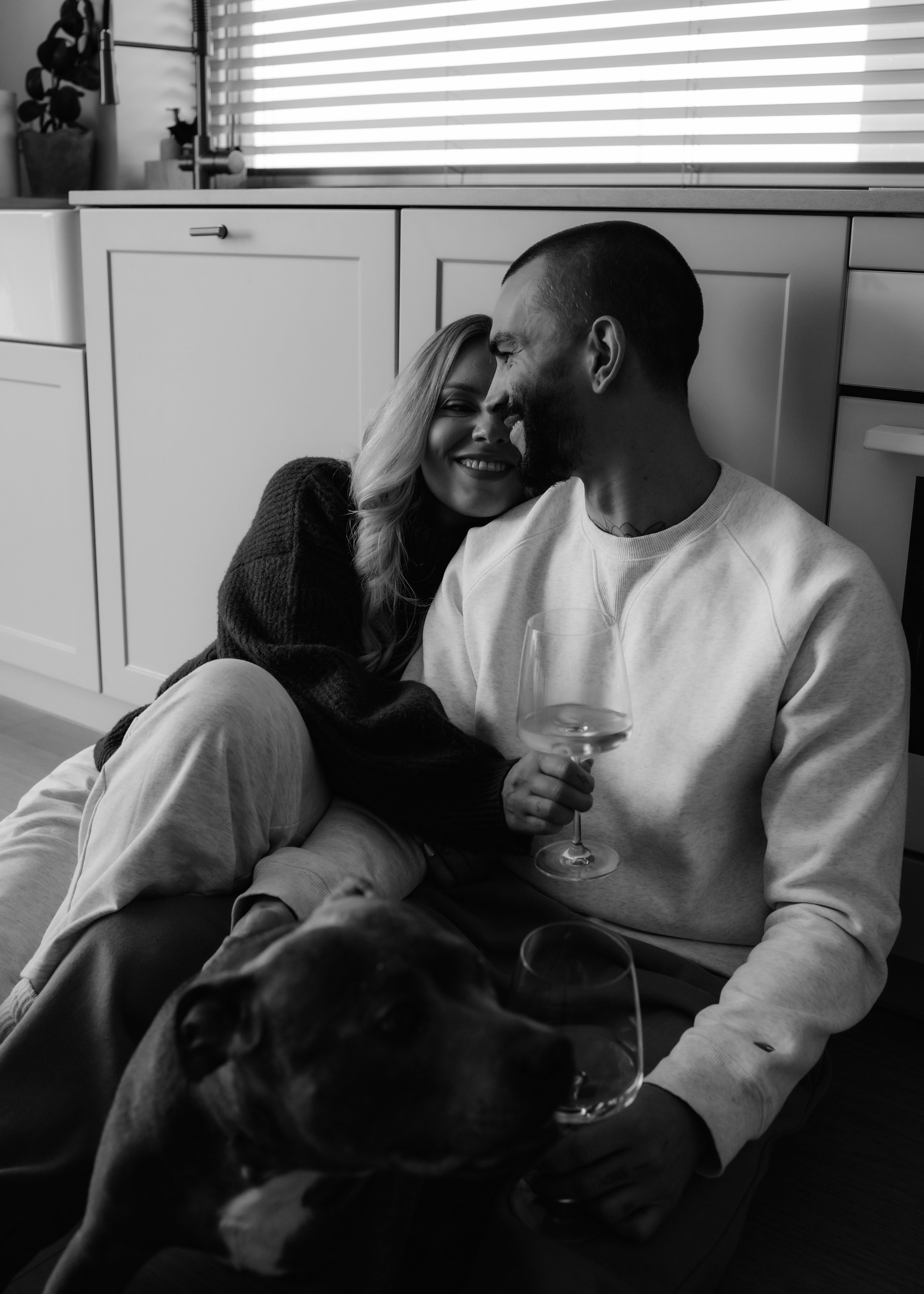 A black and white photo of a smiling woman and a man with a beard, sitting closely on the floor inside a home, with the woman leaning into the man's face. The woman holds a glass of white wine, and a dog is in the foreground. The background features 