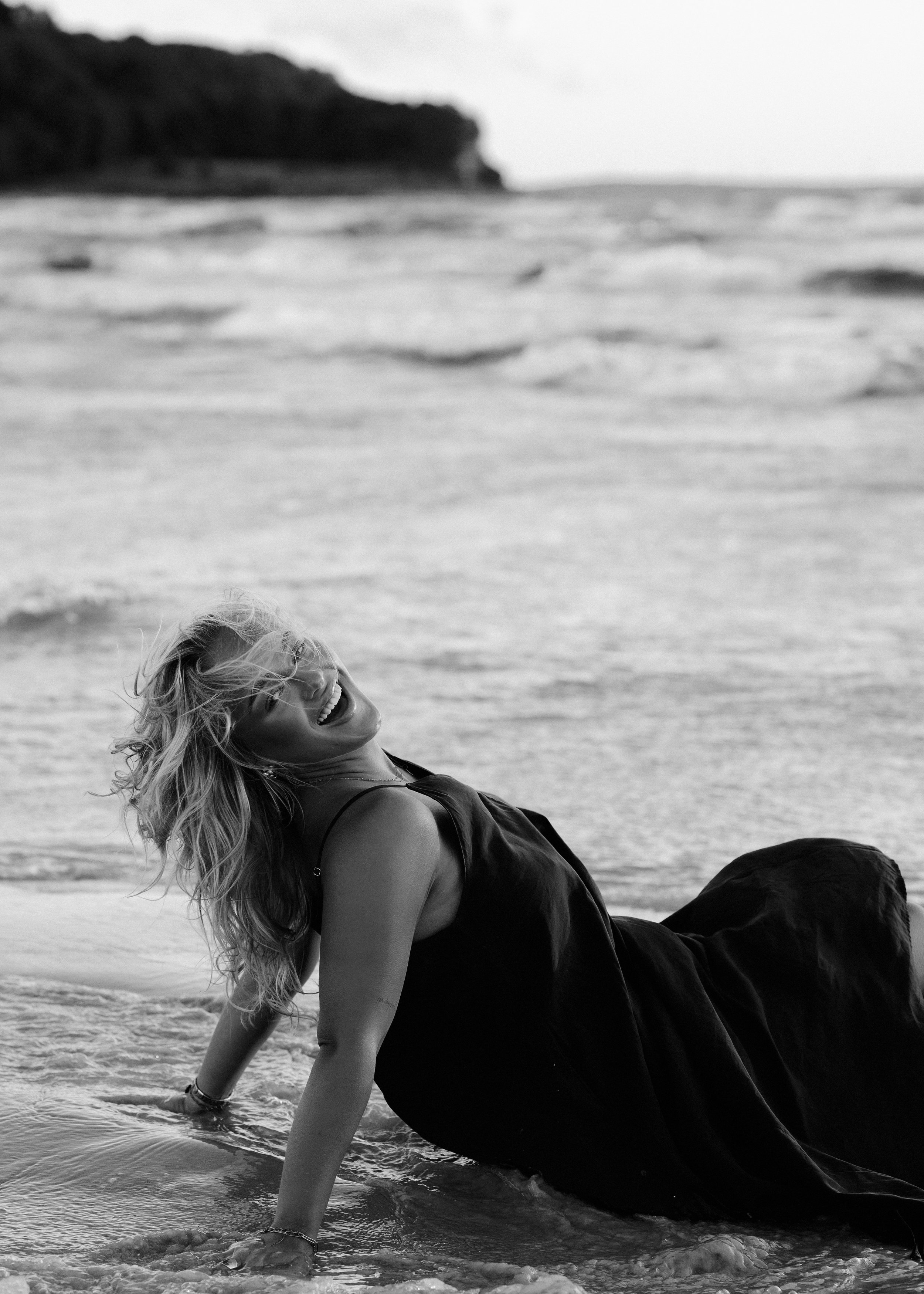 A woman with long hair laughing while leaning on the shoreline with ocean waves in the background.