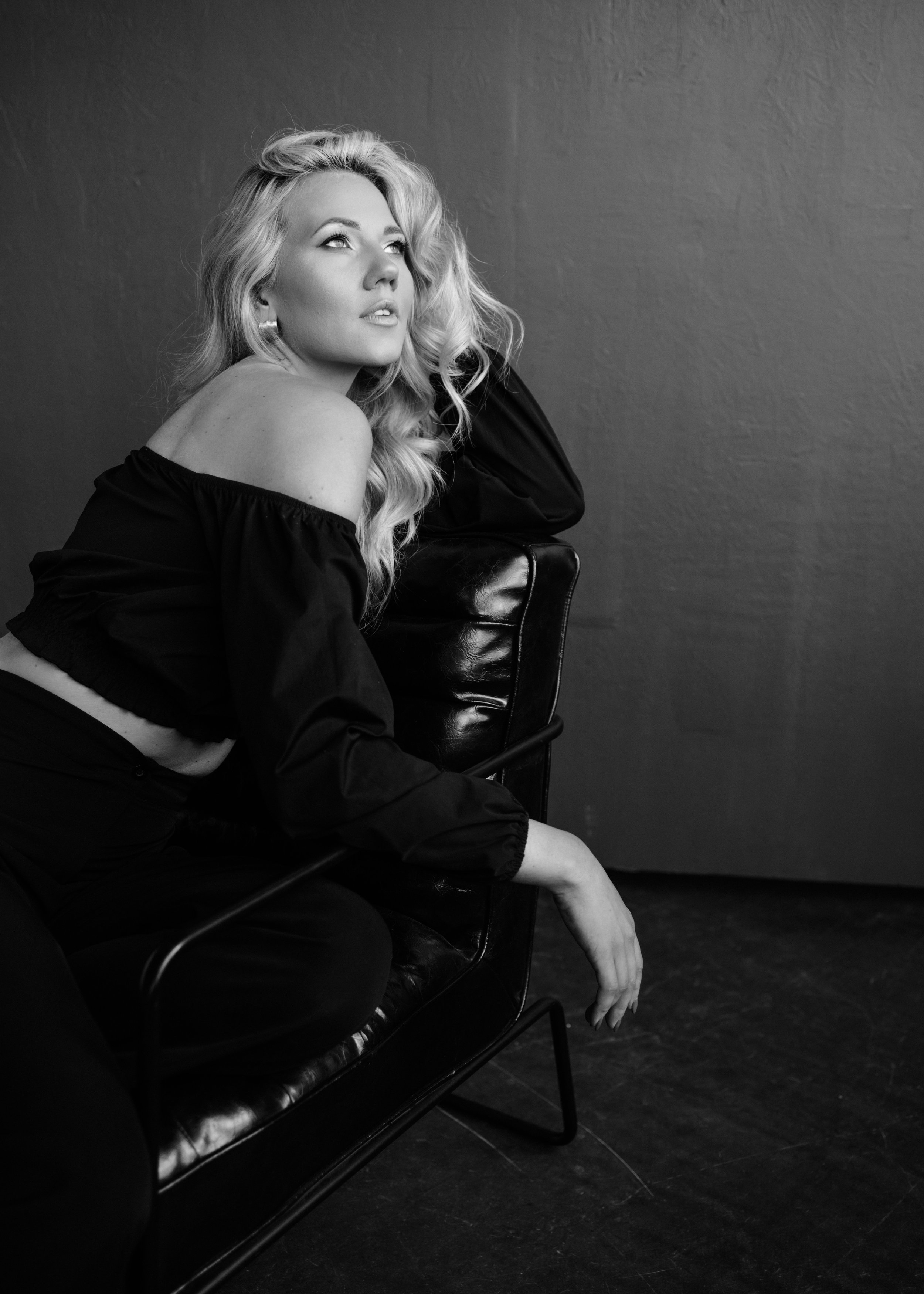 Black and white photo of a woman with long, wavy blonde hair posing next to a wall, resting her head on her hand, wearing an off-shoulder black top, sitting on a chair.