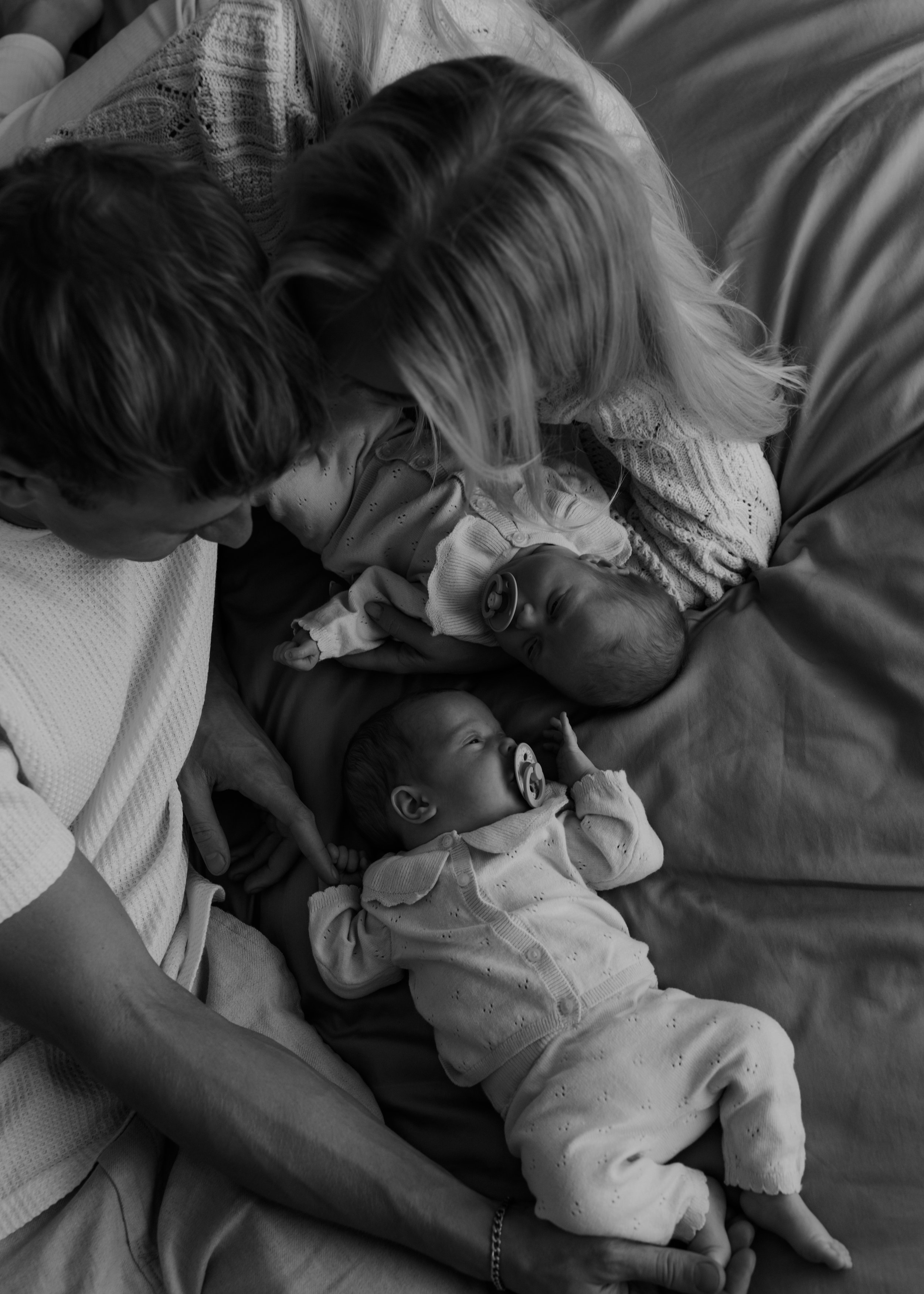 A black and white photo of two adults and two infants lying together on a bed or couch. The adults are leaning over the babies, who are looking at the adults and each other, one baby with a pacifier.