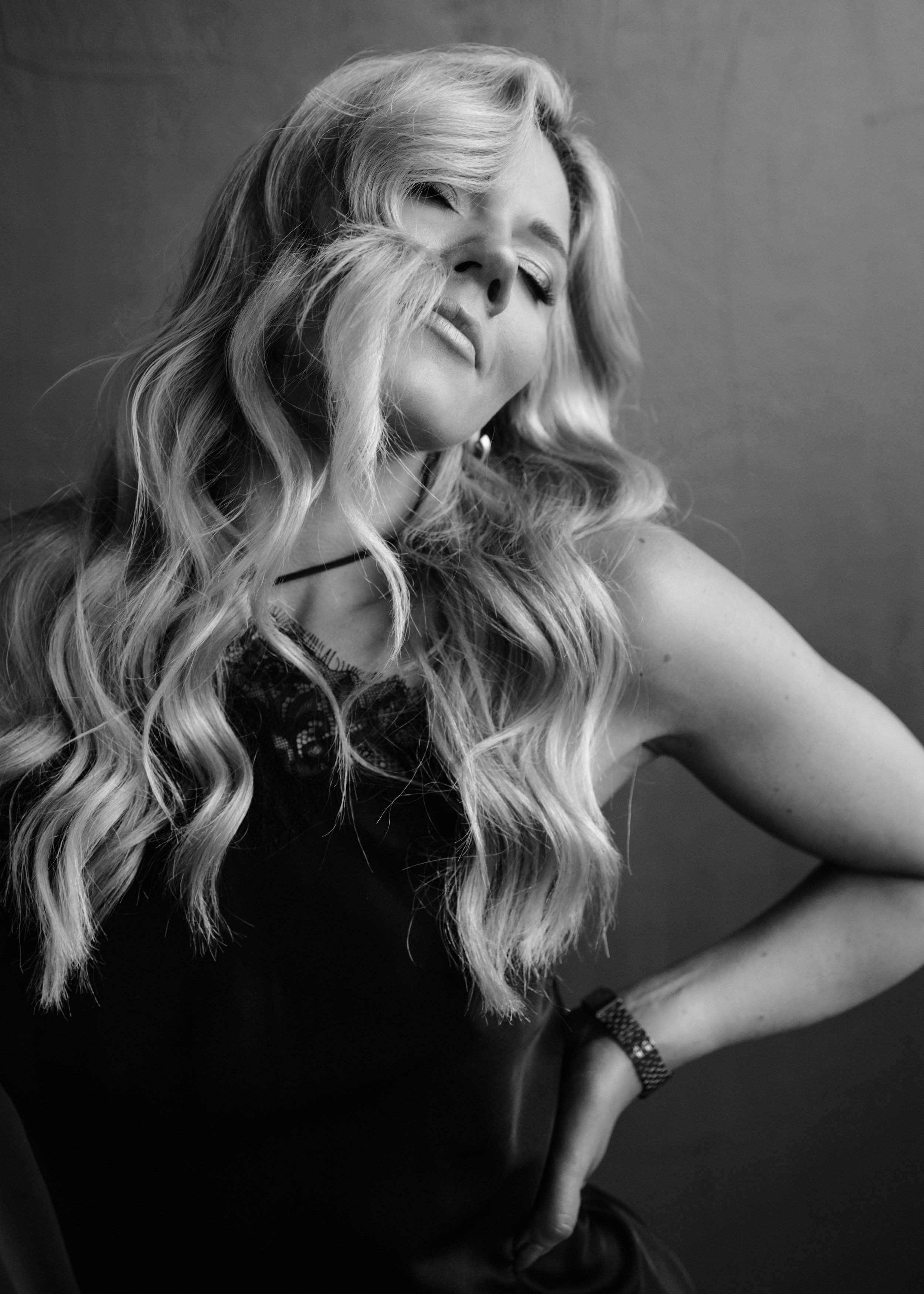 Black and white portrait of a woman with long, wavy blond hair, partially covering her face, posing with her eyes closed and her chin tilted, wearing a dark sleeveless top and a wristwatch.