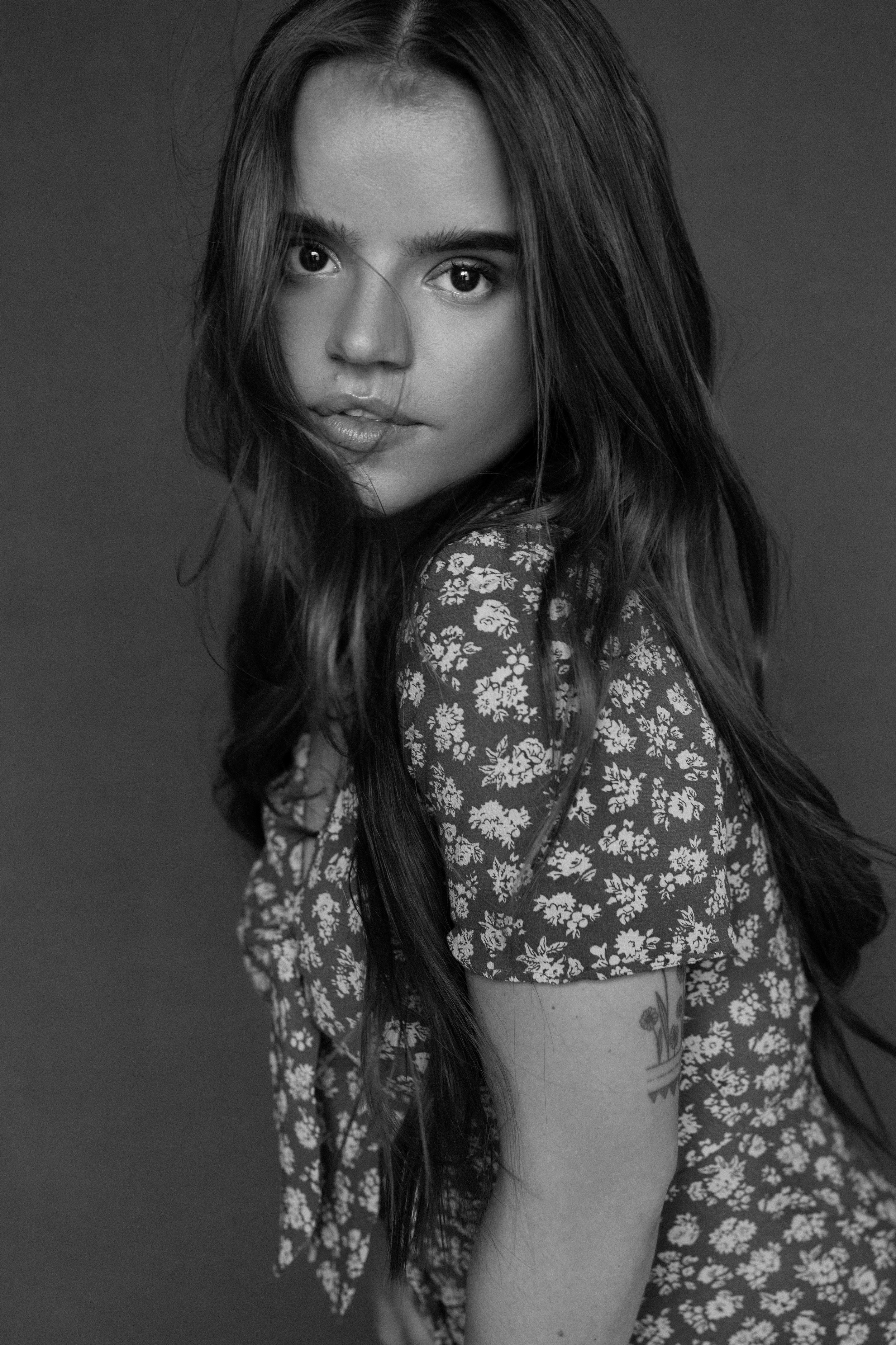 Black and white photo of a woman with long hair, wearing a floral dress, looking at the camera with an intense expression.