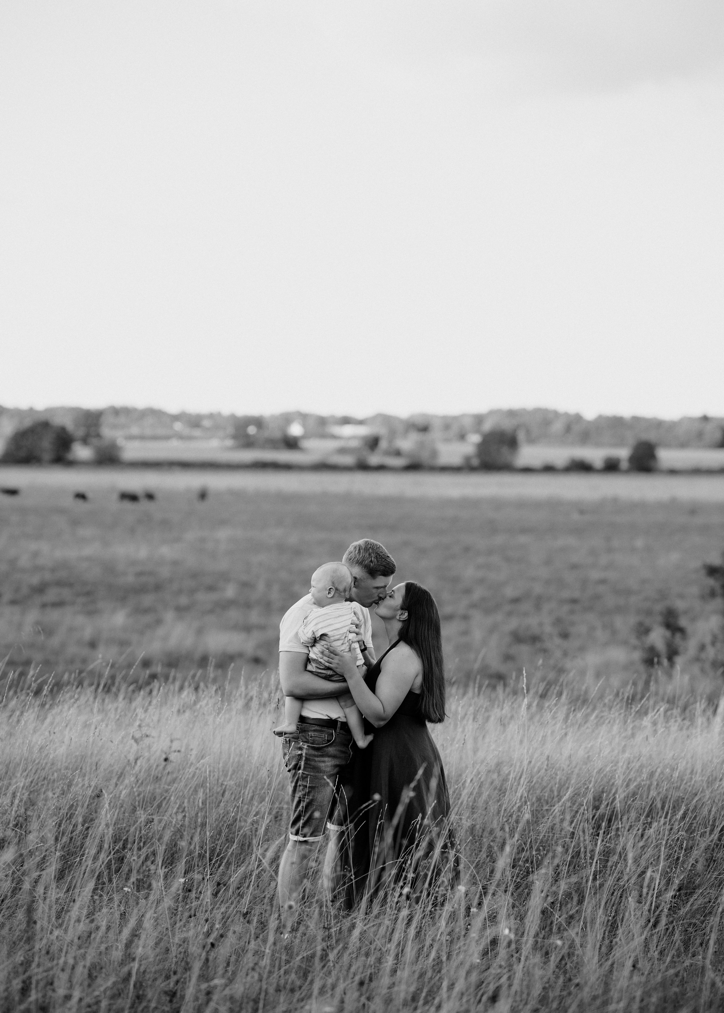 A black-and-white photo of a family in a grassy field, with a man, woman, and young child; the man and woman are kissing while the man holds the child.