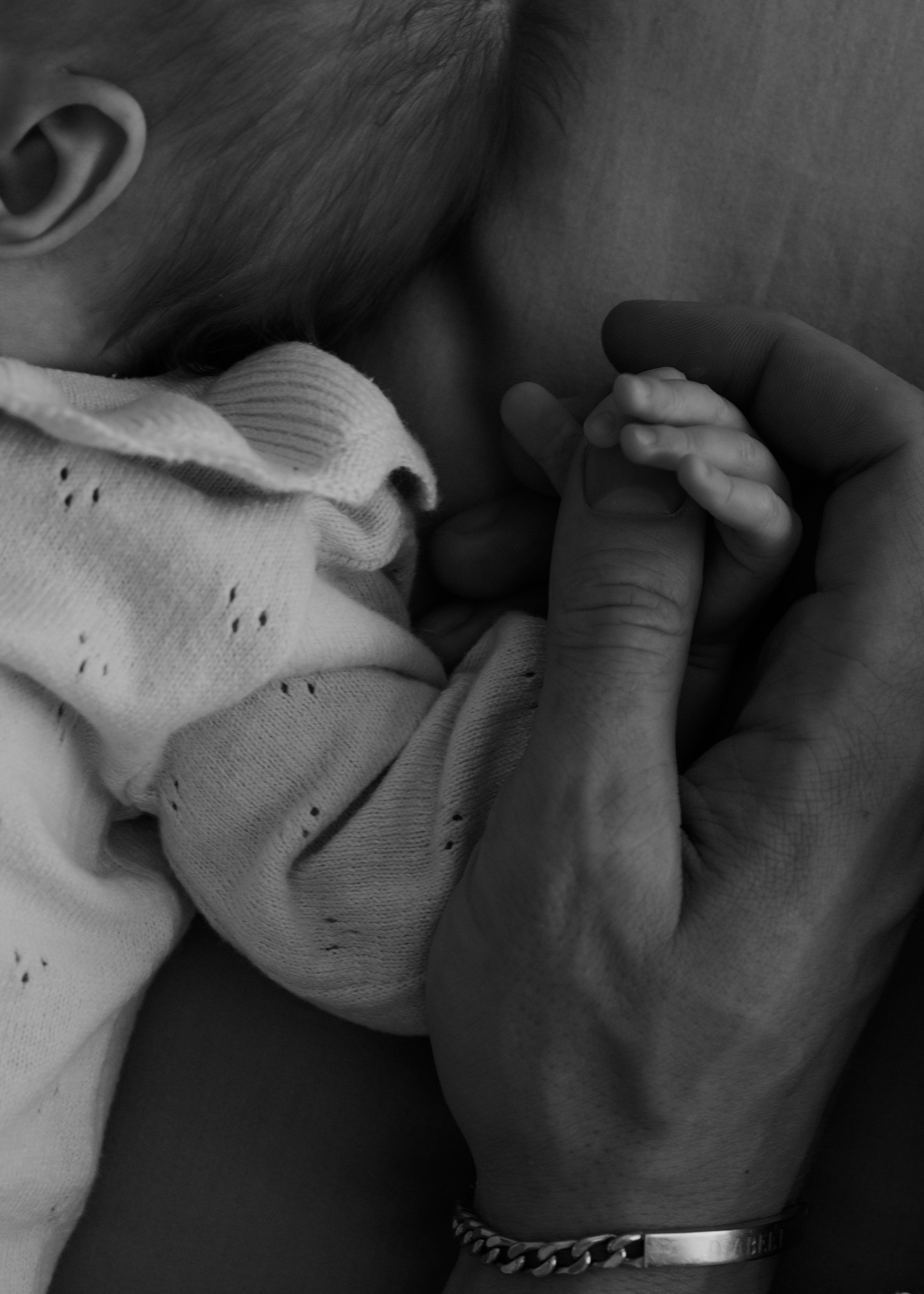 Close-up black and white photo of a baby holding an adult's finger, with an adult wearing a wristwatch. The baby is wearing a sweater with holes.