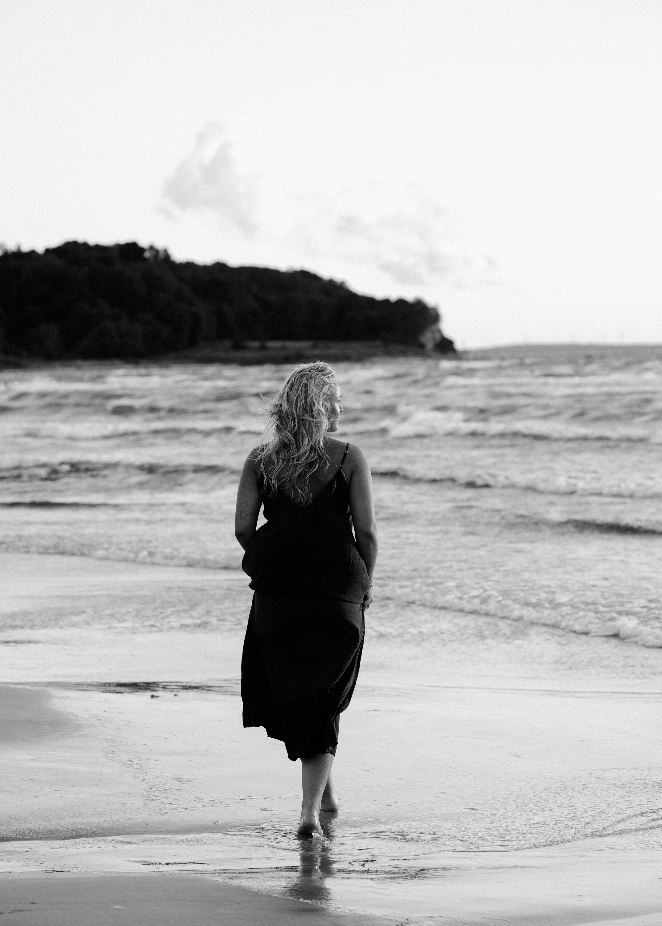 A woman with long, wavy hair standing in the shallow water at the beach, facing away from the camera, with a distant shoreline and hills in the background, in black and white.
