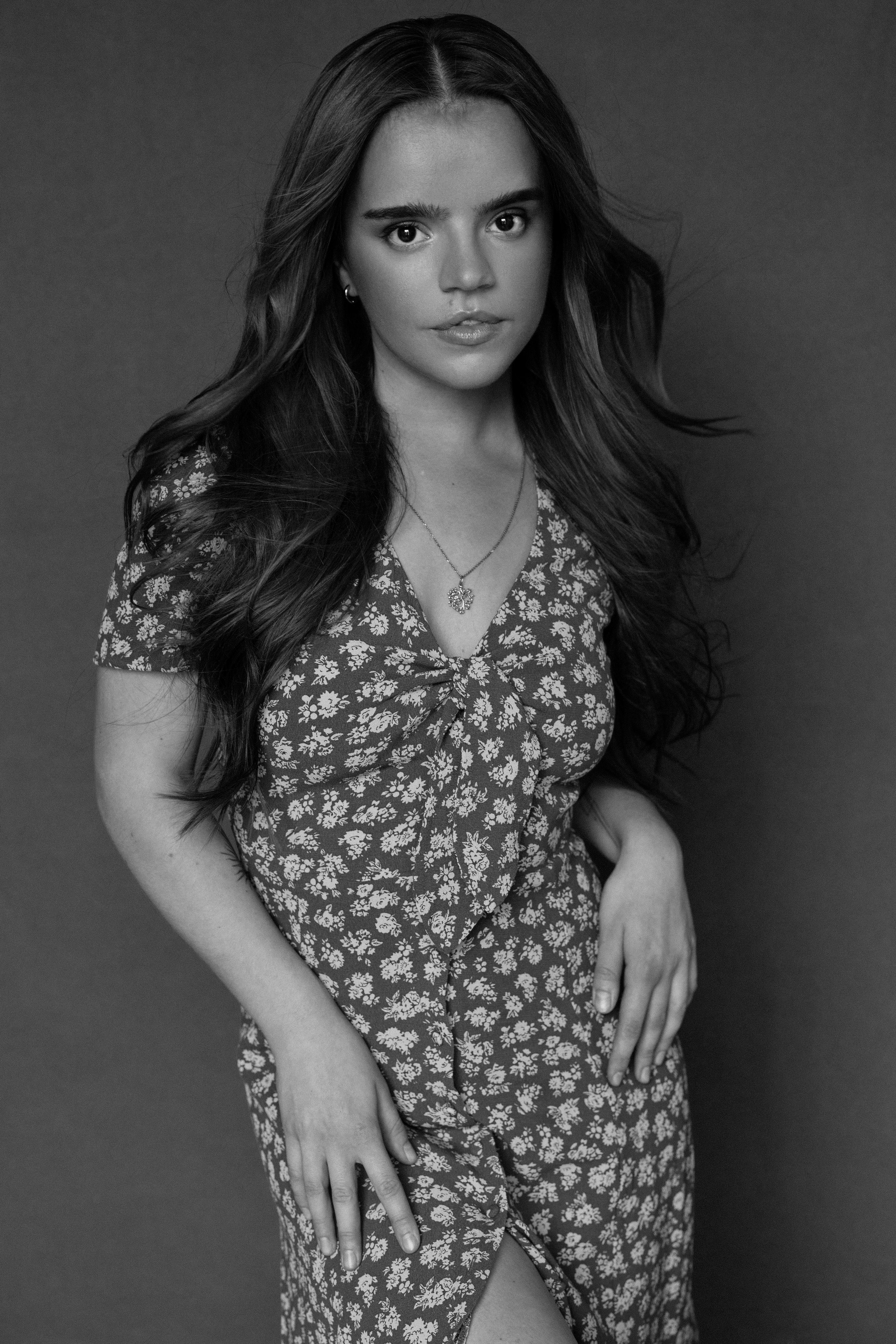 Black and white portrait of a young woman with long hair, wearing a floral dress and a necklace, posing with her hands resting on her lap.