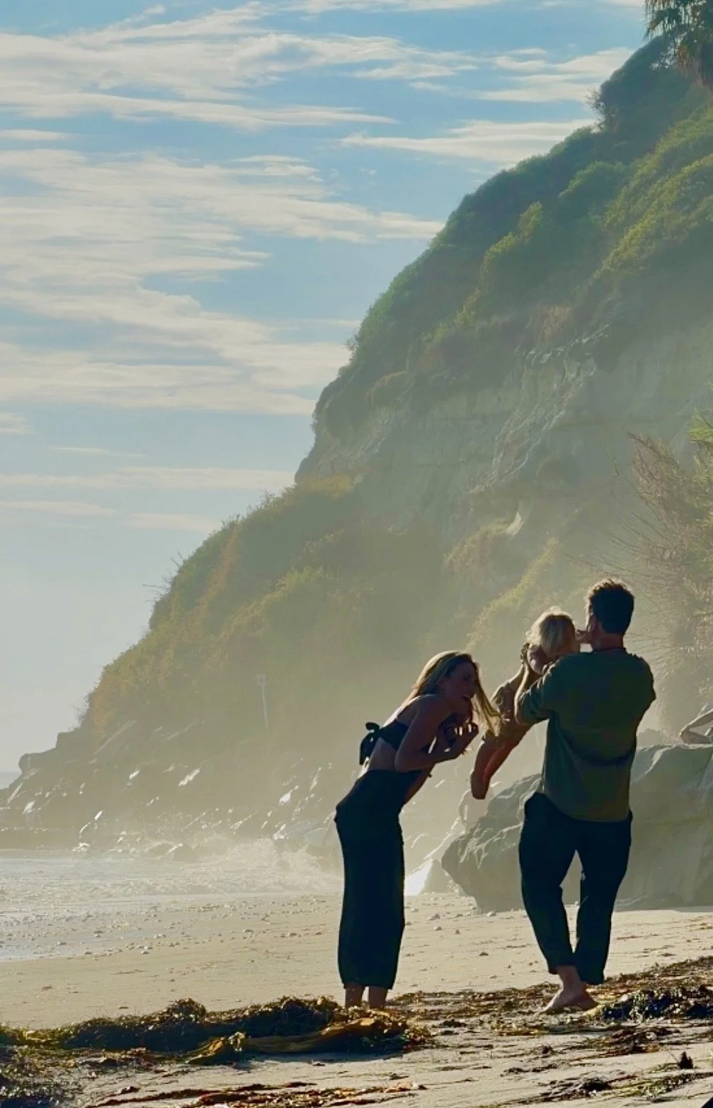 Three people on a beach with cliffs in the background; one person is holding a child, while another is leaning in and smiling.