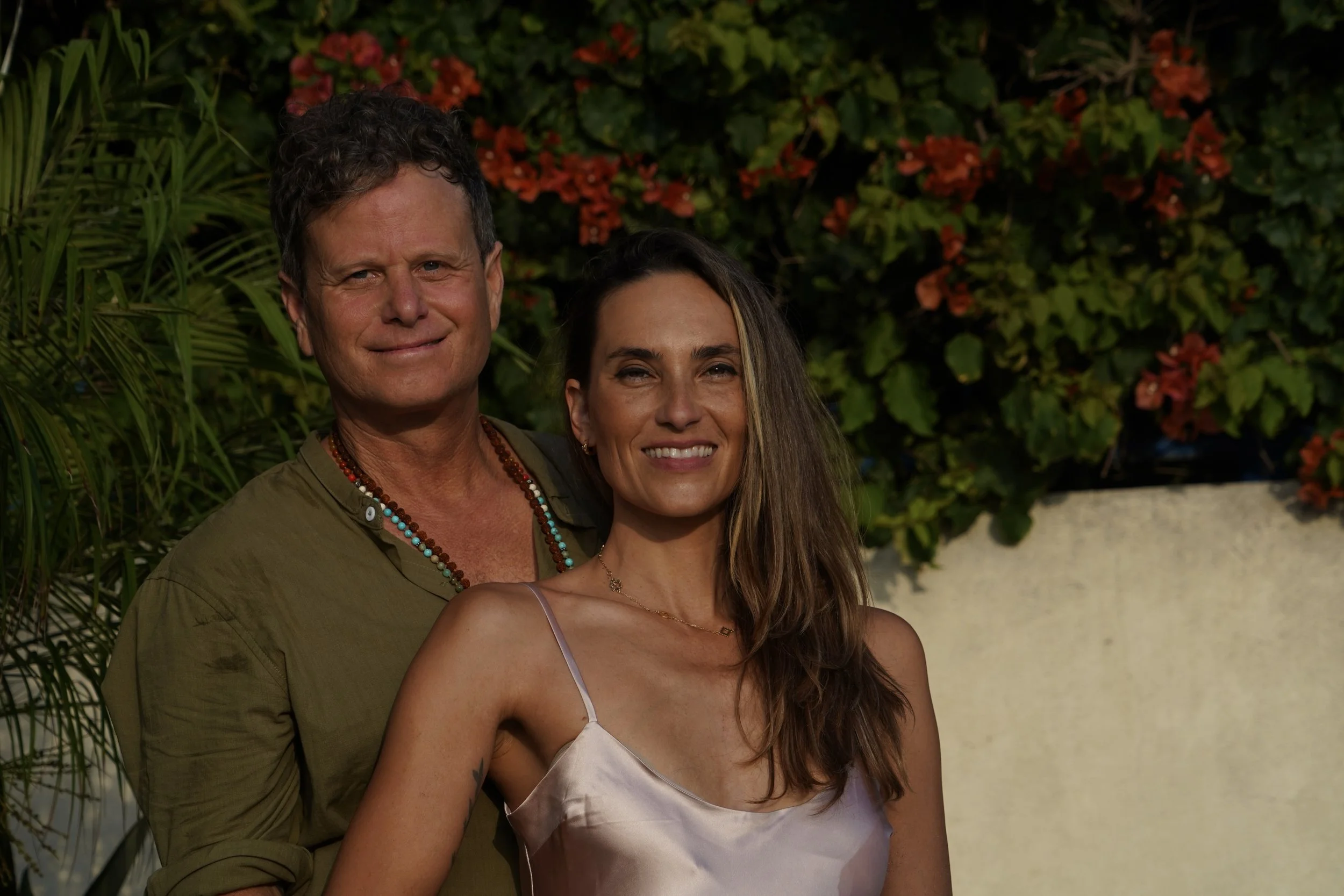 A man and a woman standing close together outdoors with green foliage and red flowers in the background, smiling at the camera.
