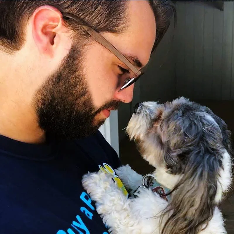 A man with glasses and a beard is touching foreheads with a small, gray and white puppy.