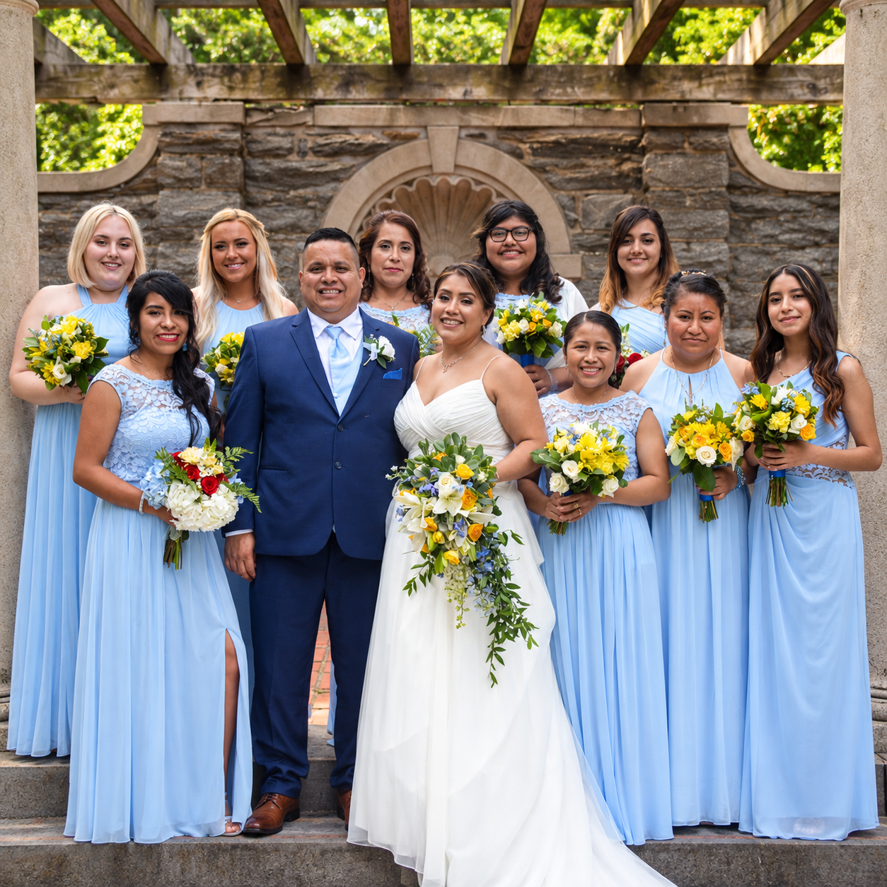 Wedding photography by David Scott Weitz of a wedding party with the bride, groom, and nine bridesmaids standing outdoors against a stone wall with greenery overhead in Elkins Park, Pennsylvania. 