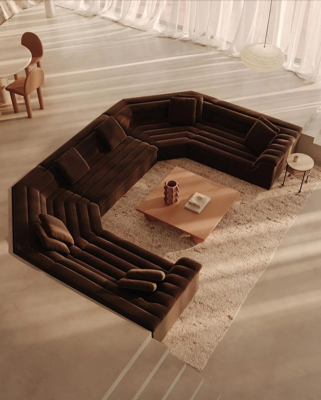 A spacious living room with a large, dark brown sectional sofa on a beige area rug, a wooden coffee table with decorative items, and a small side table in the corner. Light curtains and natural light fill the background.