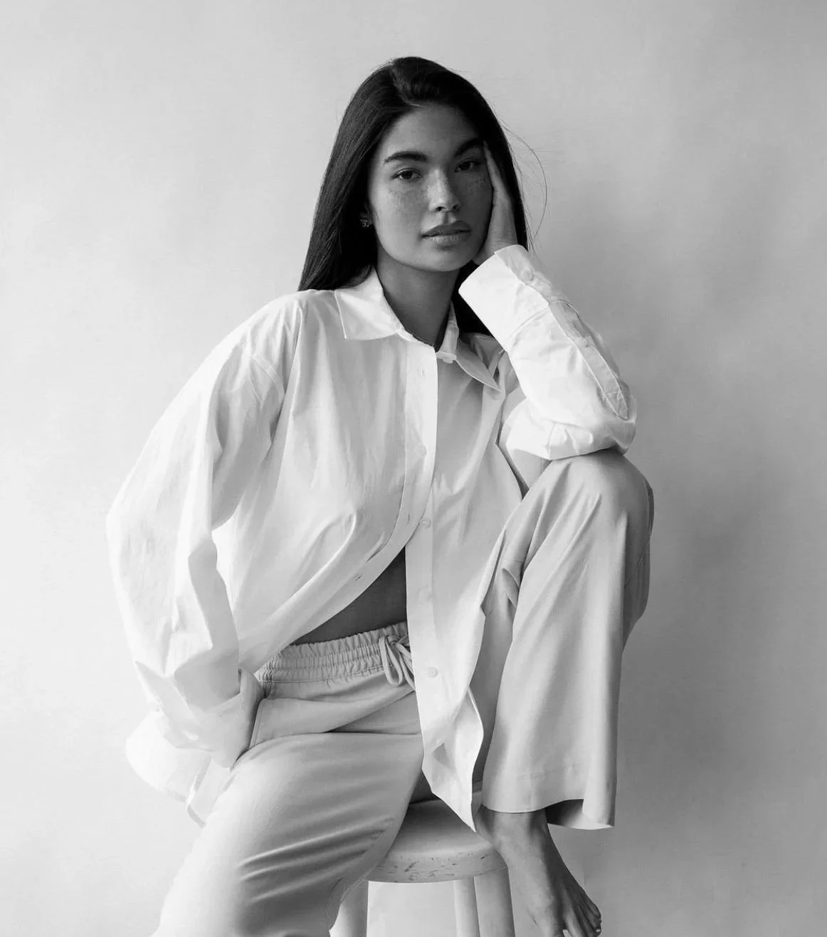 A black and white photo of a young woman with long dark hair, sitting on a stool against a plain wall, wearing loose white clothing, with one hand resting on her cheek and a relaxed expression.