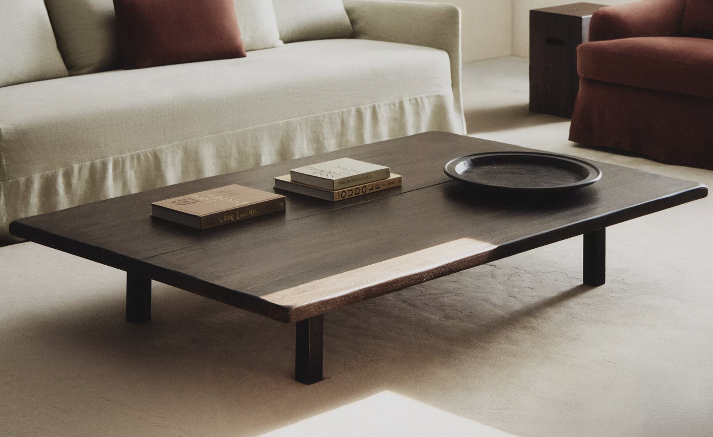 A wooden coffee table with three books and a black tray on top, in a living room with beige carpet, a light-colored sofa with pillows, and a red armchair in the background.