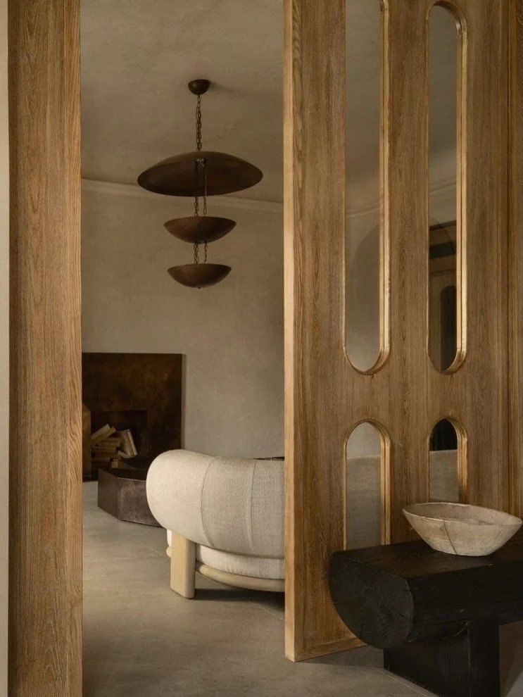 Interior view of a living room with a modern chandelier, a beige curved sofa, a wooden room divider with oval cutouts, and a dark side table with a stone bowl on top.