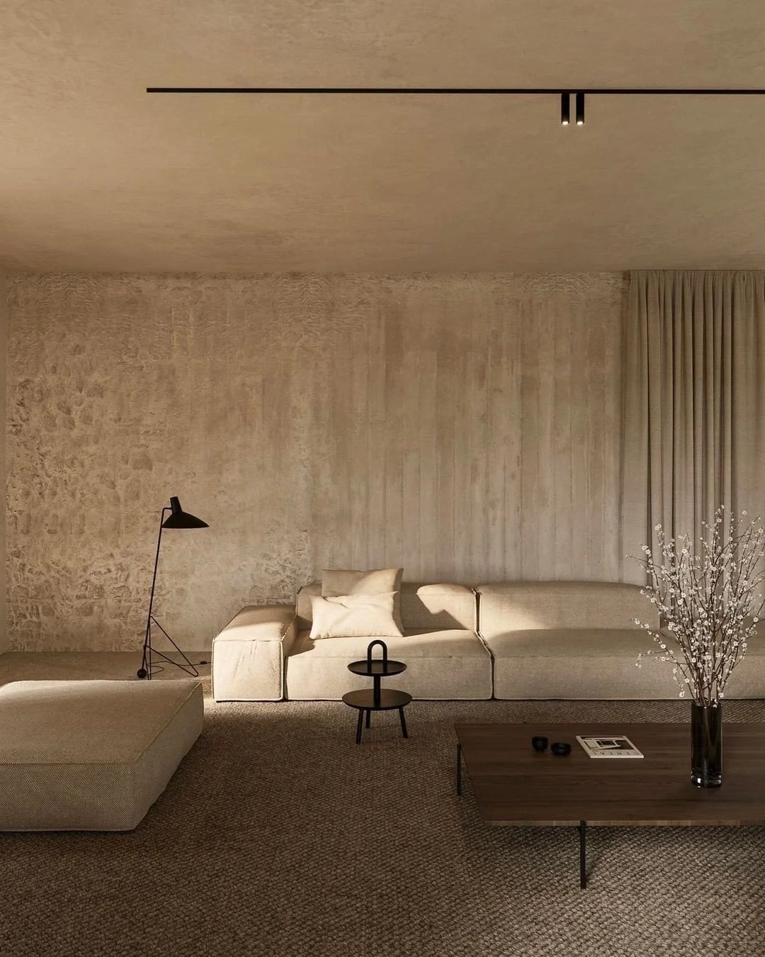 A minimalist living room with beige sofas, a coffee table, a flower vase with white flowers, a black floor lamp, and wall-mounted ceiling lights.