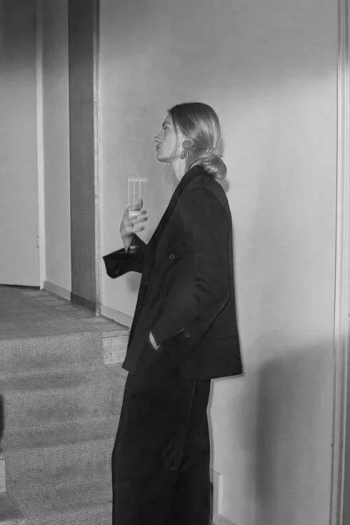 A woman in a black blazer and pants holding a glass, leaning against a wall in a modern interior space.