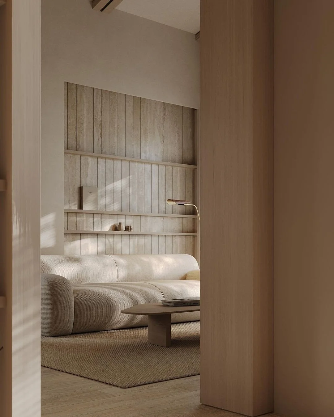 Minimalist beige living room with a beige sofa, a wooden coffee table with magazines, and a built-in wooden wall shelf with small decorative objects and a book, illuminated by natural light.