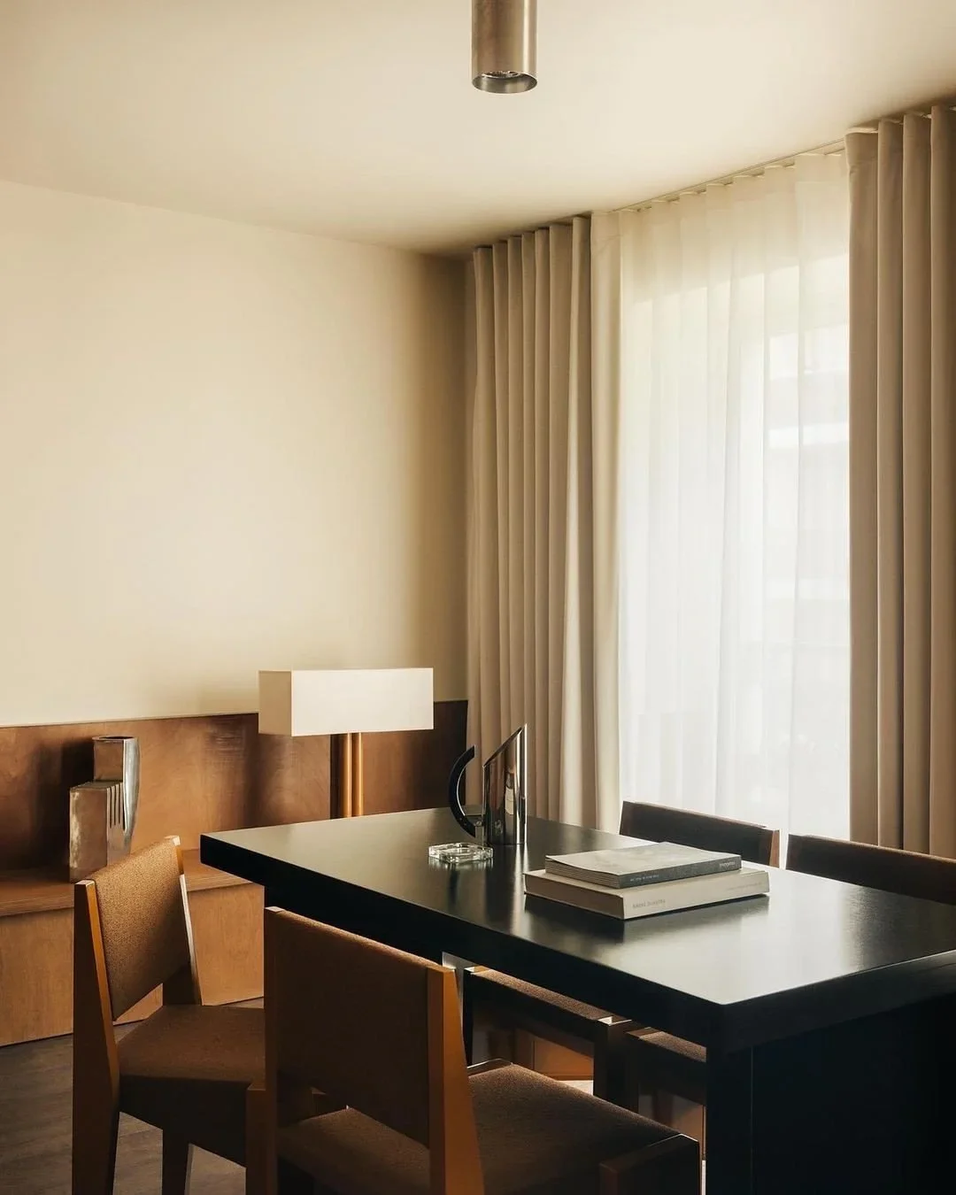 Modern dining room with black table, brown chairs, white curtains, beige walls, wooden sideboard, and a white lamp.