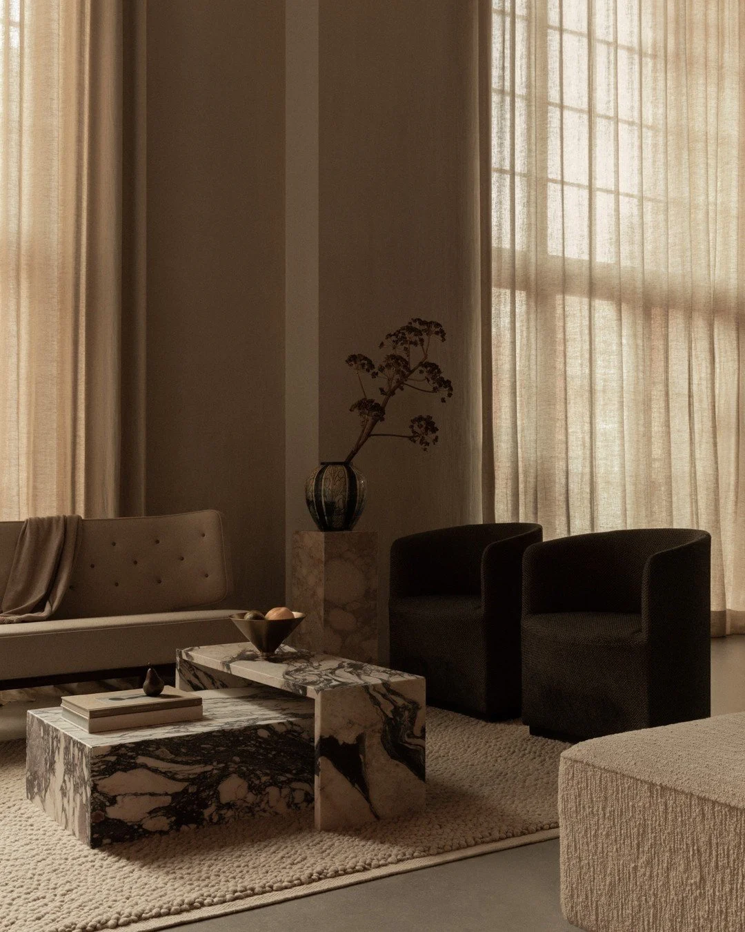 A living room with beige curtains, a beige sofa, two black armchairs, a marble coffee table, a beige textured rug, a decorative vase with dried branches, and a stack of books.