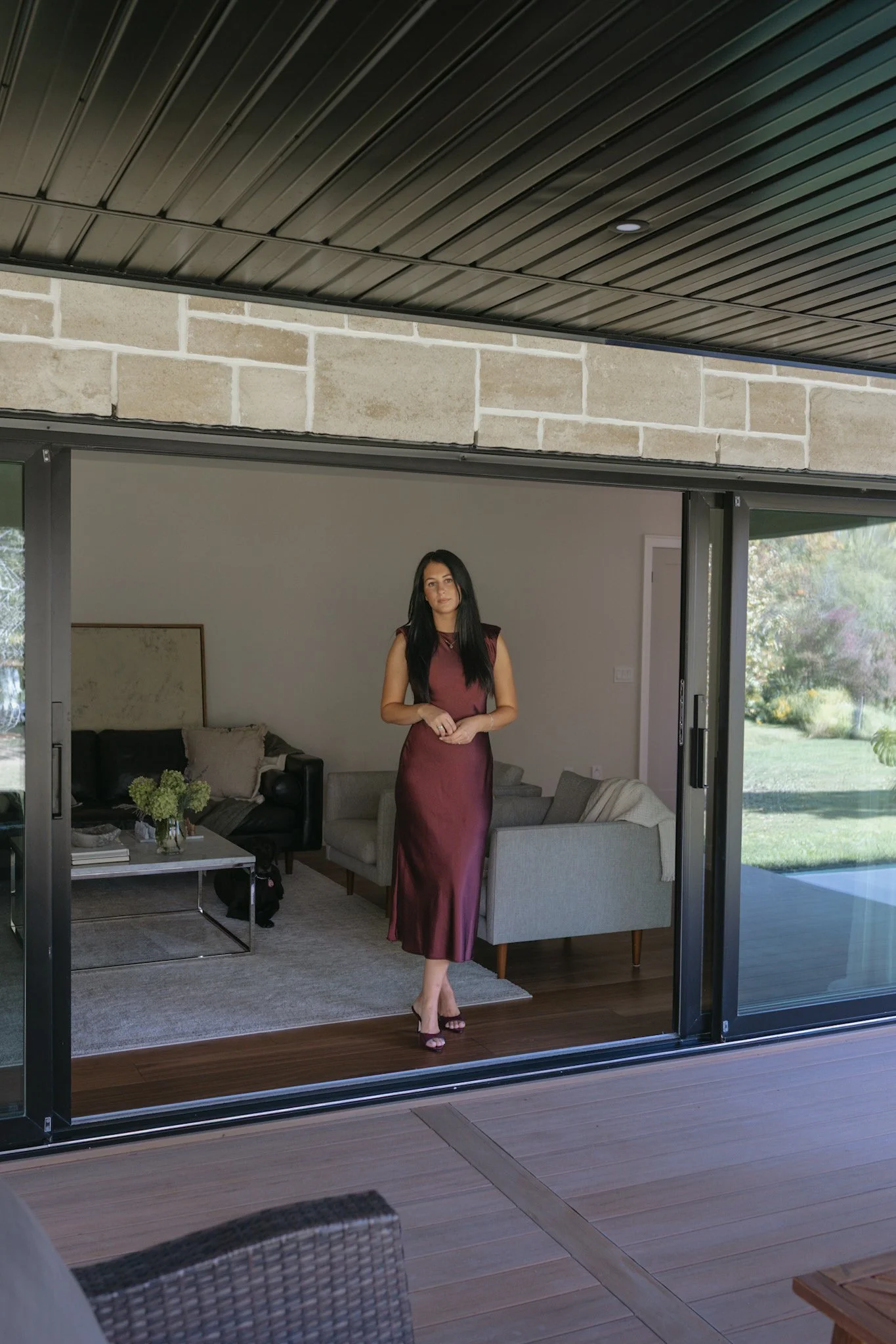 A woman in a sleeveless, burgundy satin dress with high-heeled sandals standing inside a modern living room with large glass doors opening to a backyard.